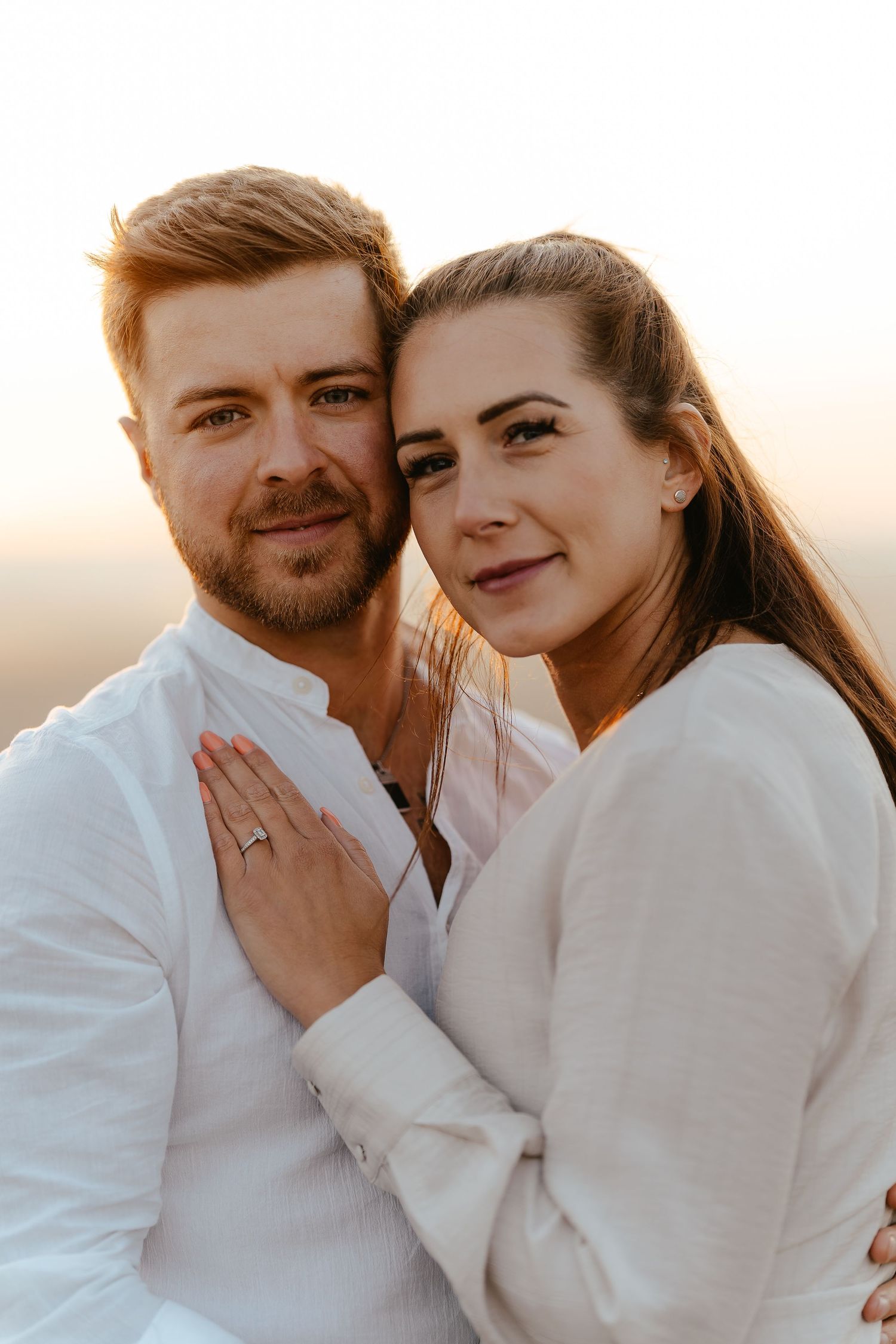 A smiling couple posing cheek to cheek at sunset, woman displaying an engagement ring on her hand.