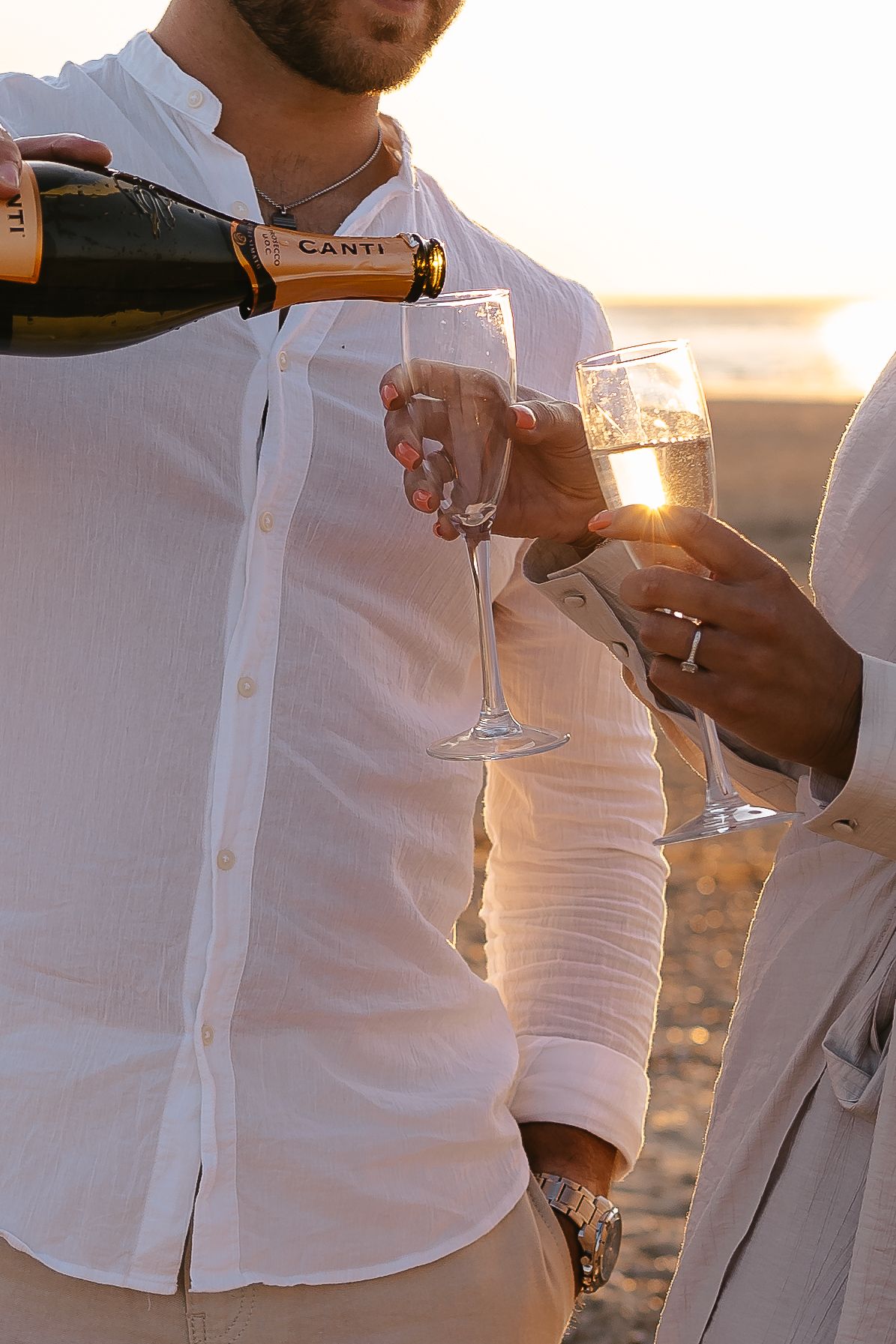 Close-up of a man pouring Canti champagne into glasses held by a couple on a beach at sunset.