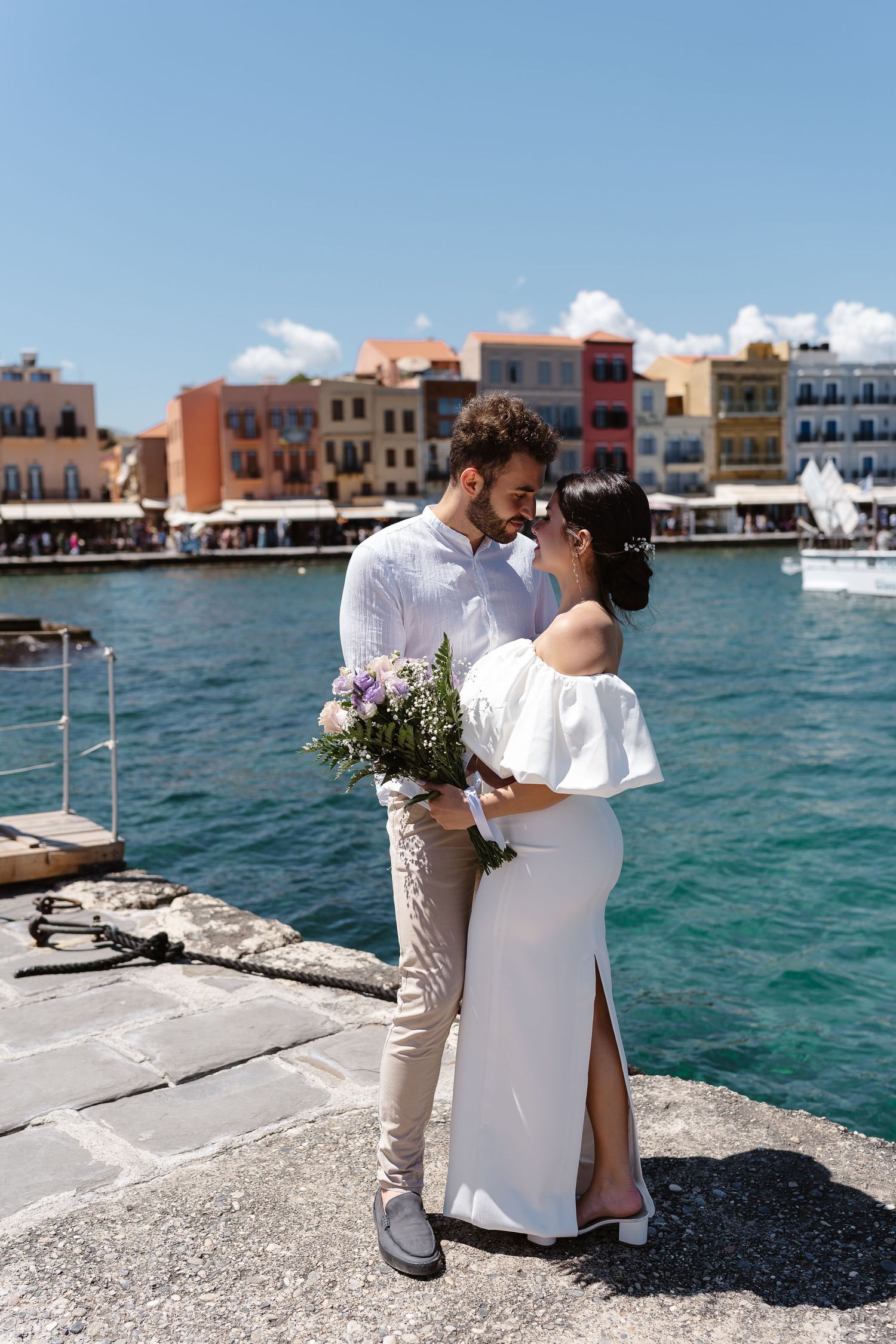 Happy couple shares a romantic moment on a Greek harbor pier, colorful Venetian buildings reflected in turquoise water behind them.