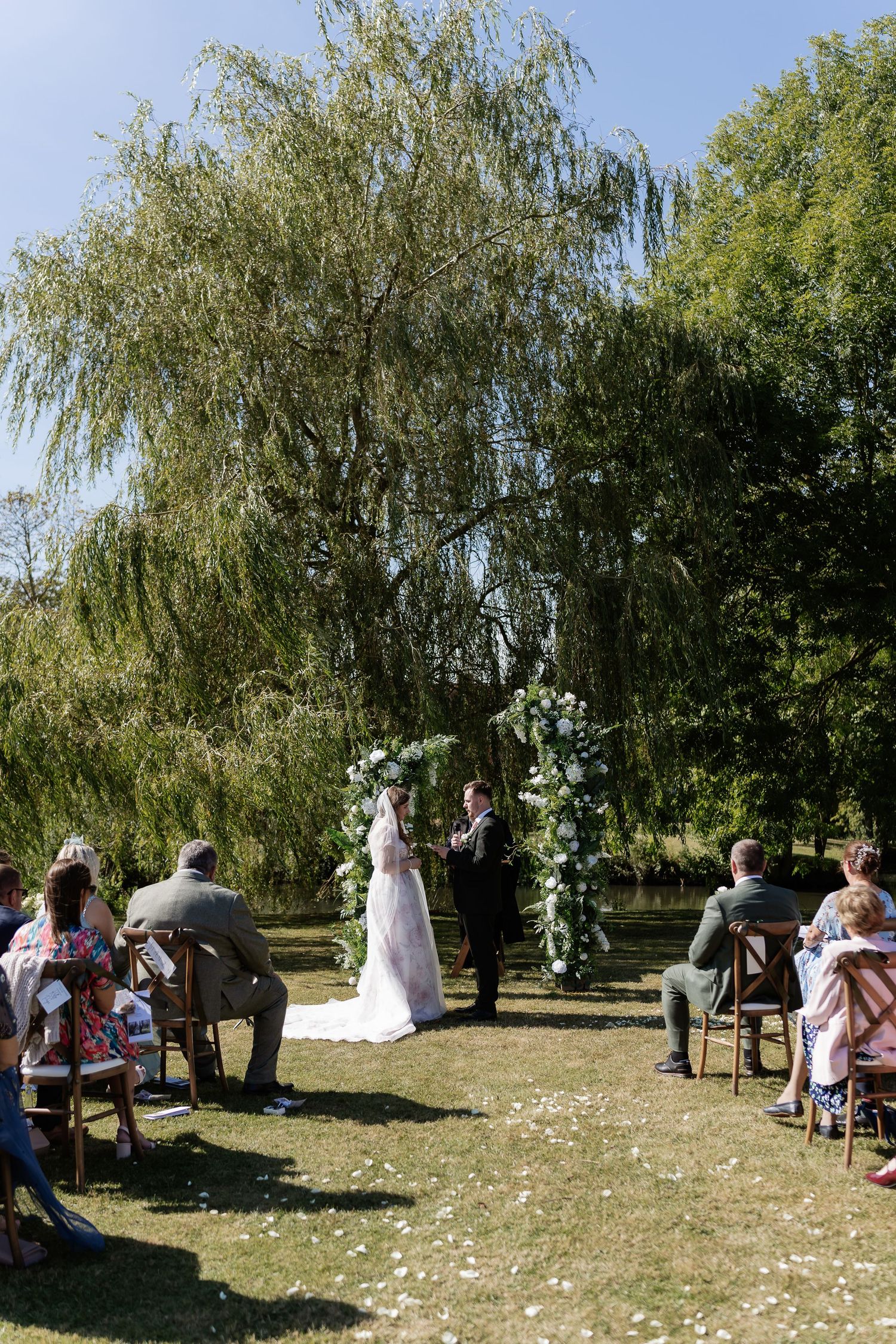 Bride and groom exchange vows under a floral arch beside a weeping willow tree at an outdoor ceremony.