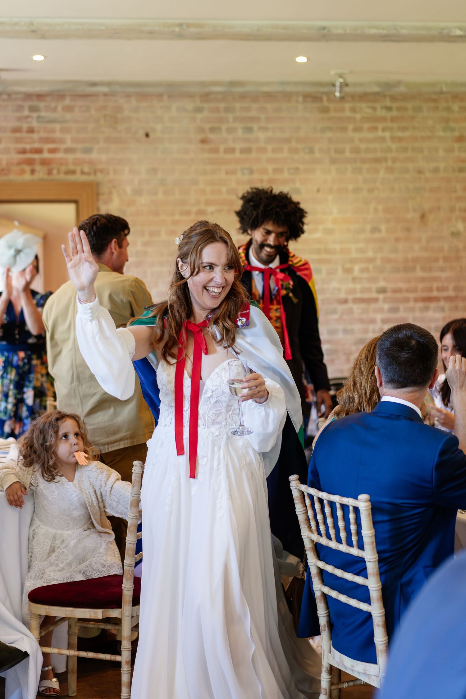 Smiling bride in white gown with red ribbon waves to guests in a rustic brick-walled wedding reception venue.