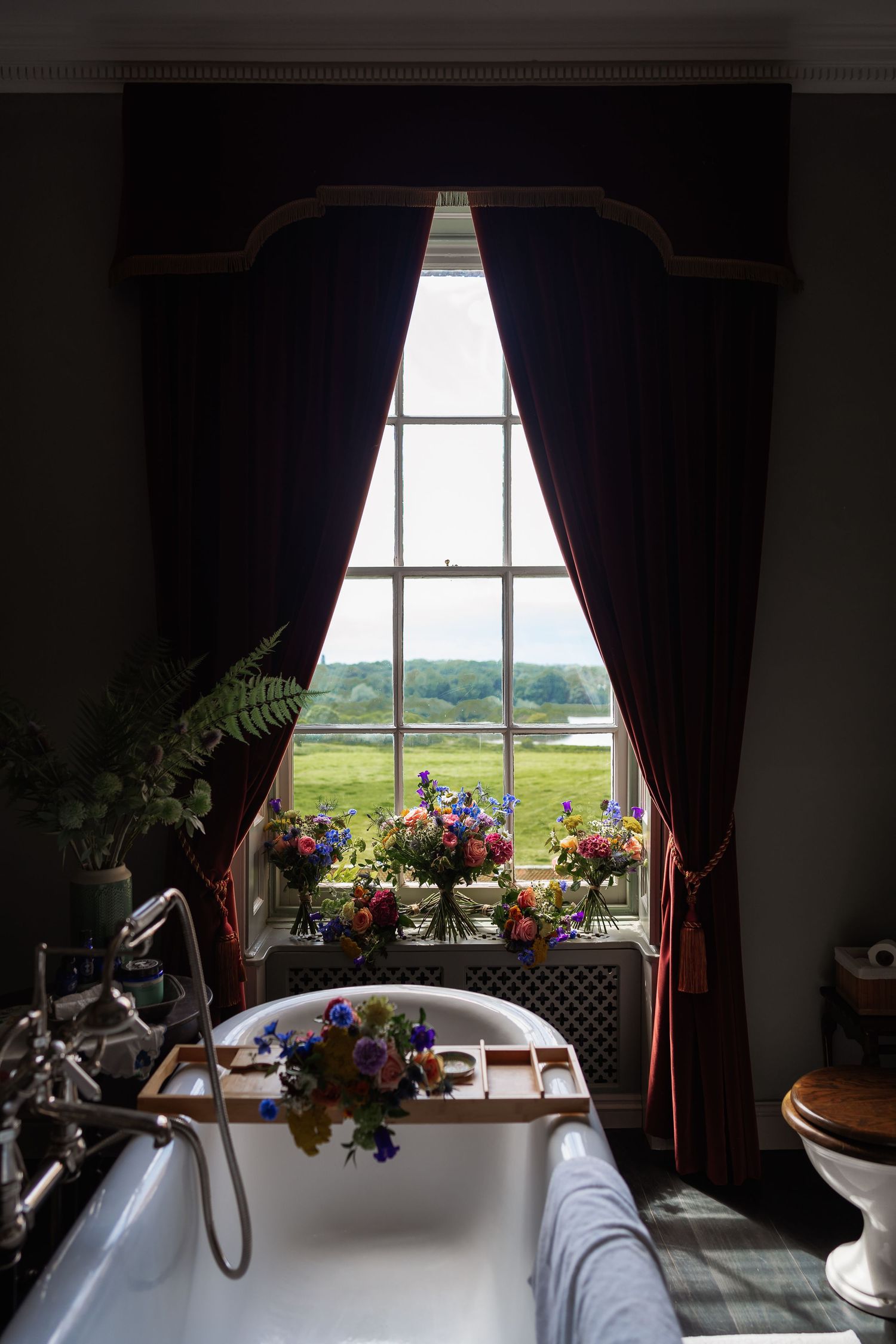 Clawfoot bathtub with flower arrangement beside tall draped window overlooking countryside.