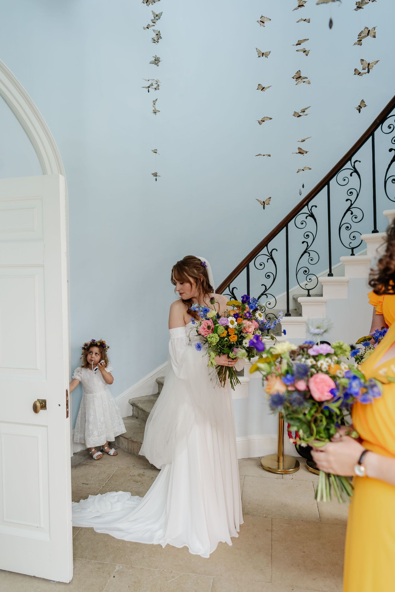 Bride in off-shoulder gown admiring colorful bouquet on ornate staircase with butterfly decorations.