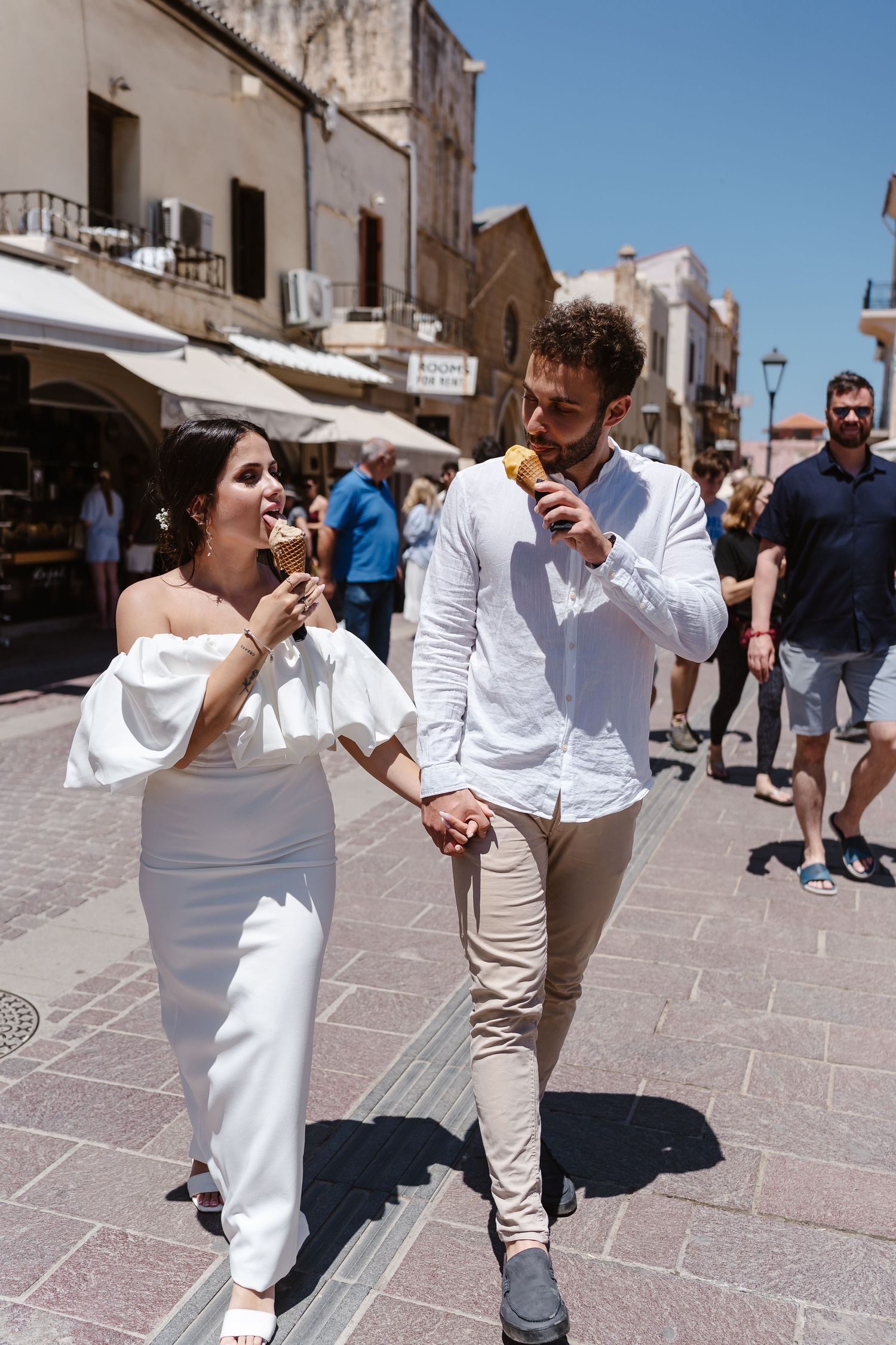 Newlywed couple holding hands and eating gelato cones while strolling through a sunny Mediterranean town square.