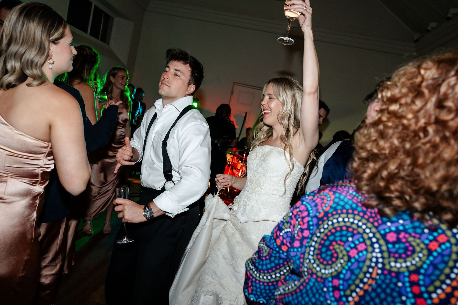 Bride and groom dance joyfully with guests on colorful lit dance floor at wedding reception.