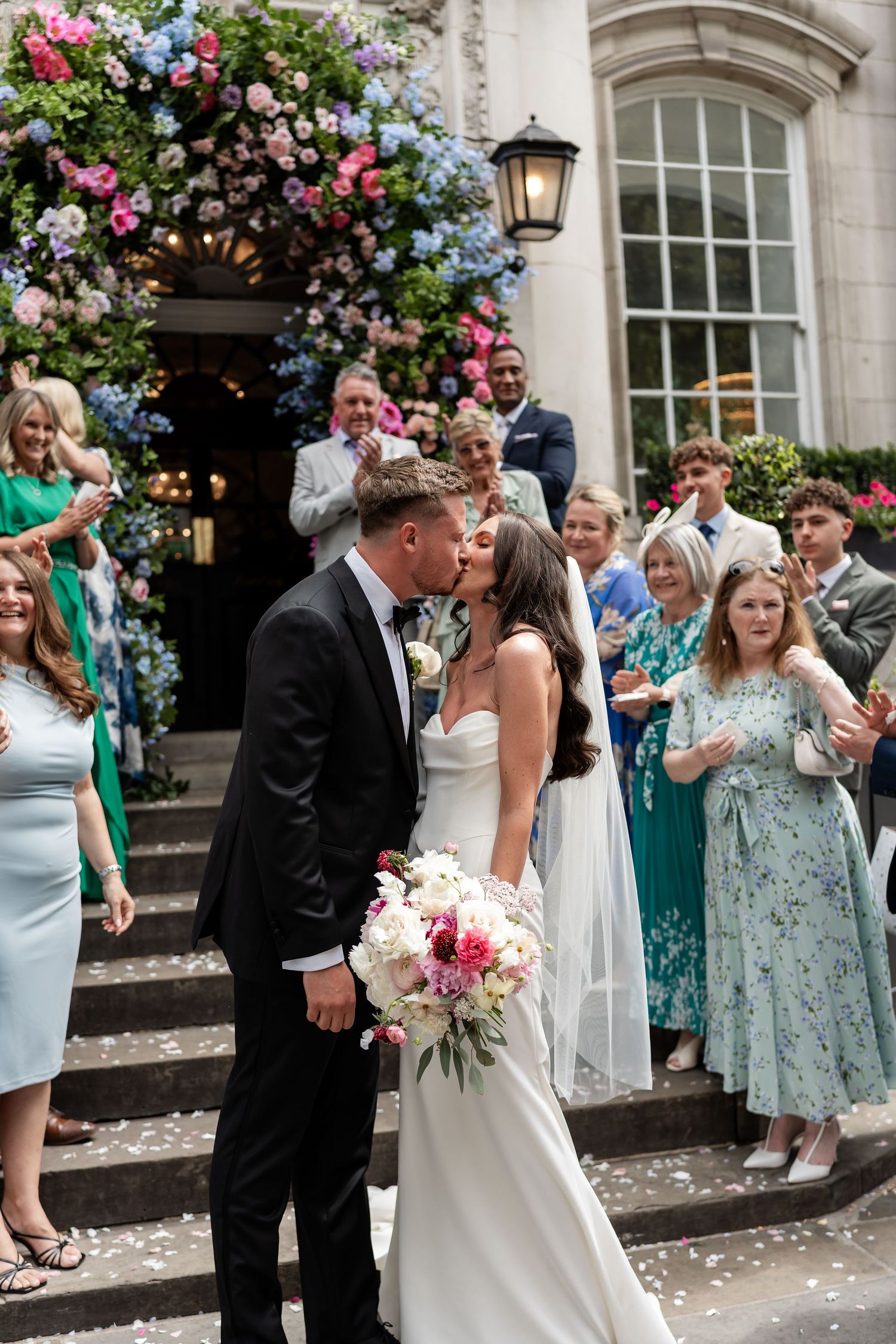 Newlyweds share kiss on steps surrounded by cheering guests and colorful floral arch entrance.