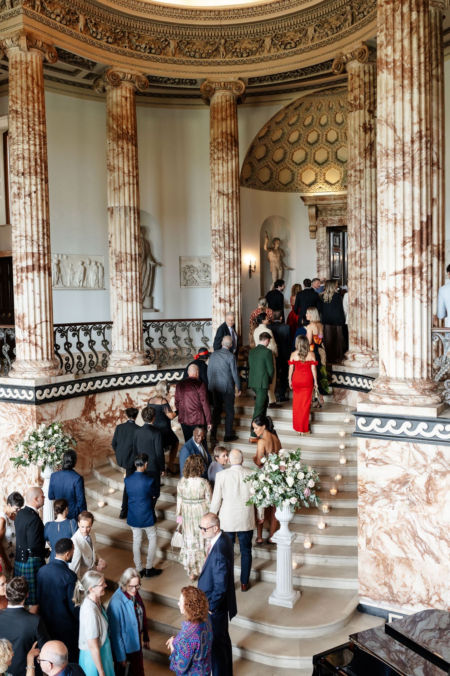 Elegant wedding guests mingle on grand marble staircase in ornate classical venue with tall columns.