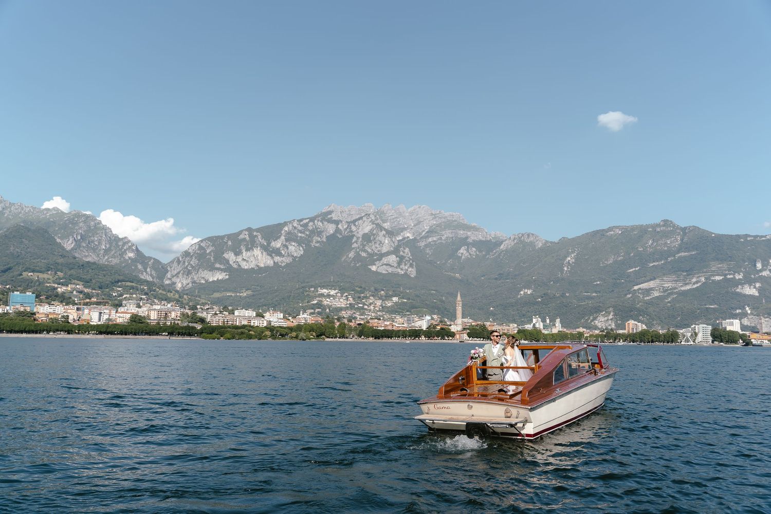 Vintage wooden boat cruises on crystal blue lake with dramatic mountain range and coastal town in background.