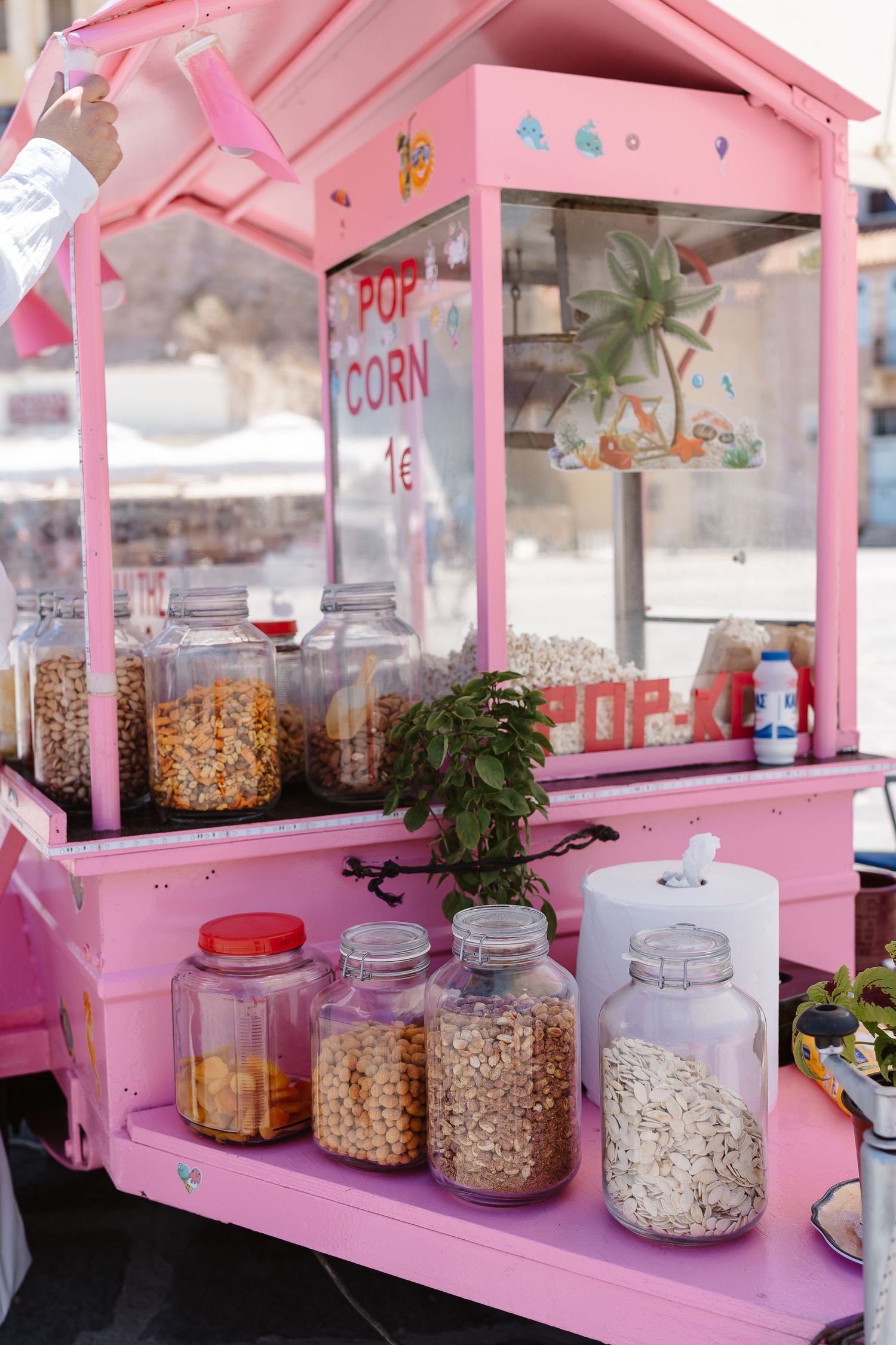 Colorful pink popcorn street cart with glass jars of snacks and a popcorn sign in a sunny plaza.