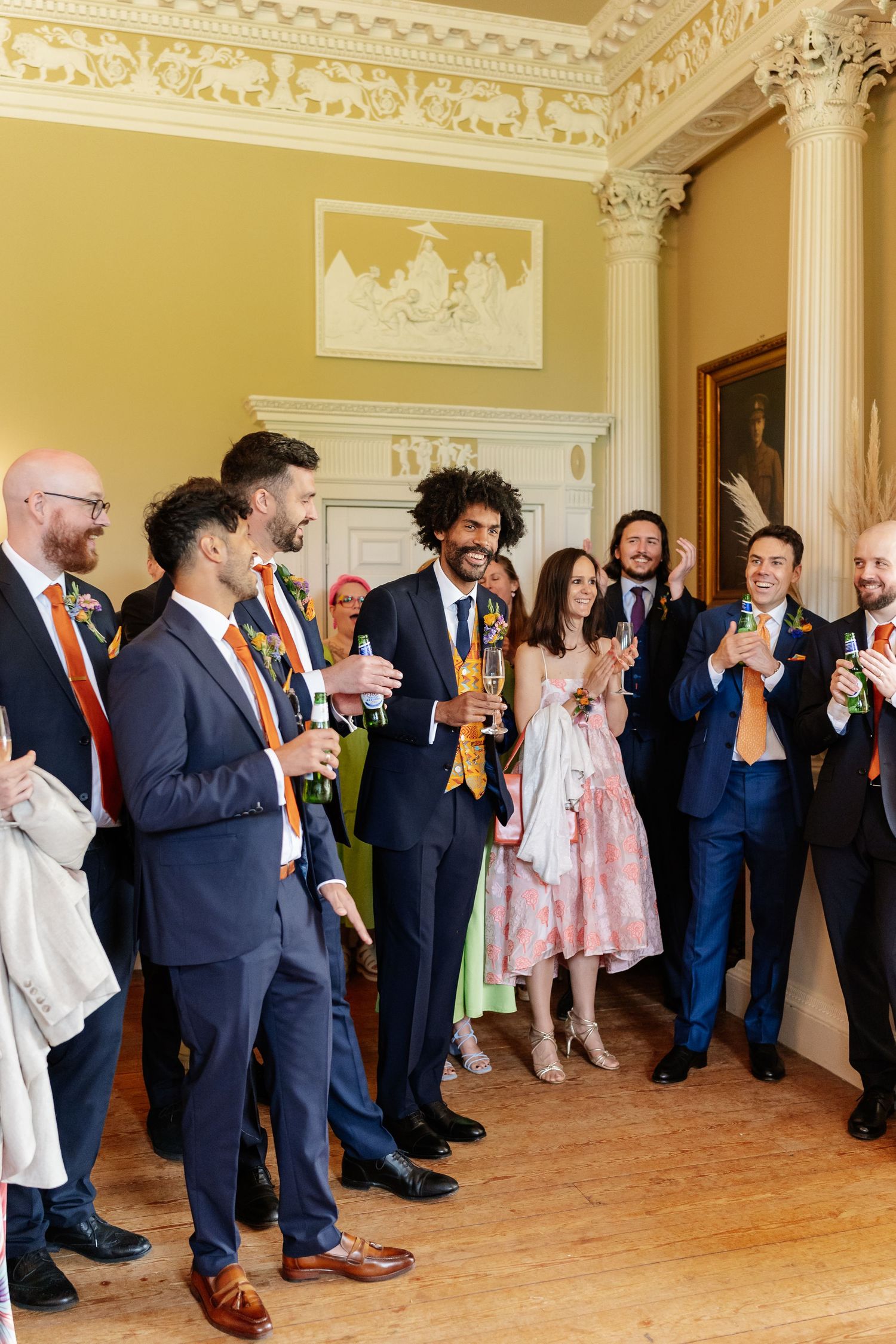 Groom celebrates with male wedding guests wearing colorful floral garlands in a grand room.