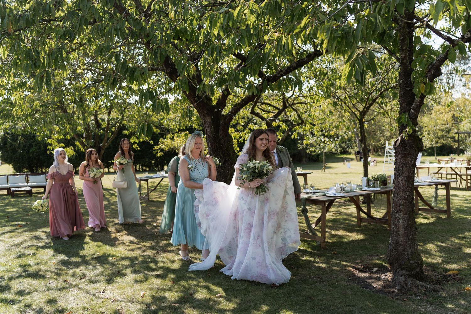 Bride in floral gown walks with bridesmaids in pink and green dresses through a sunlit garden orchard.