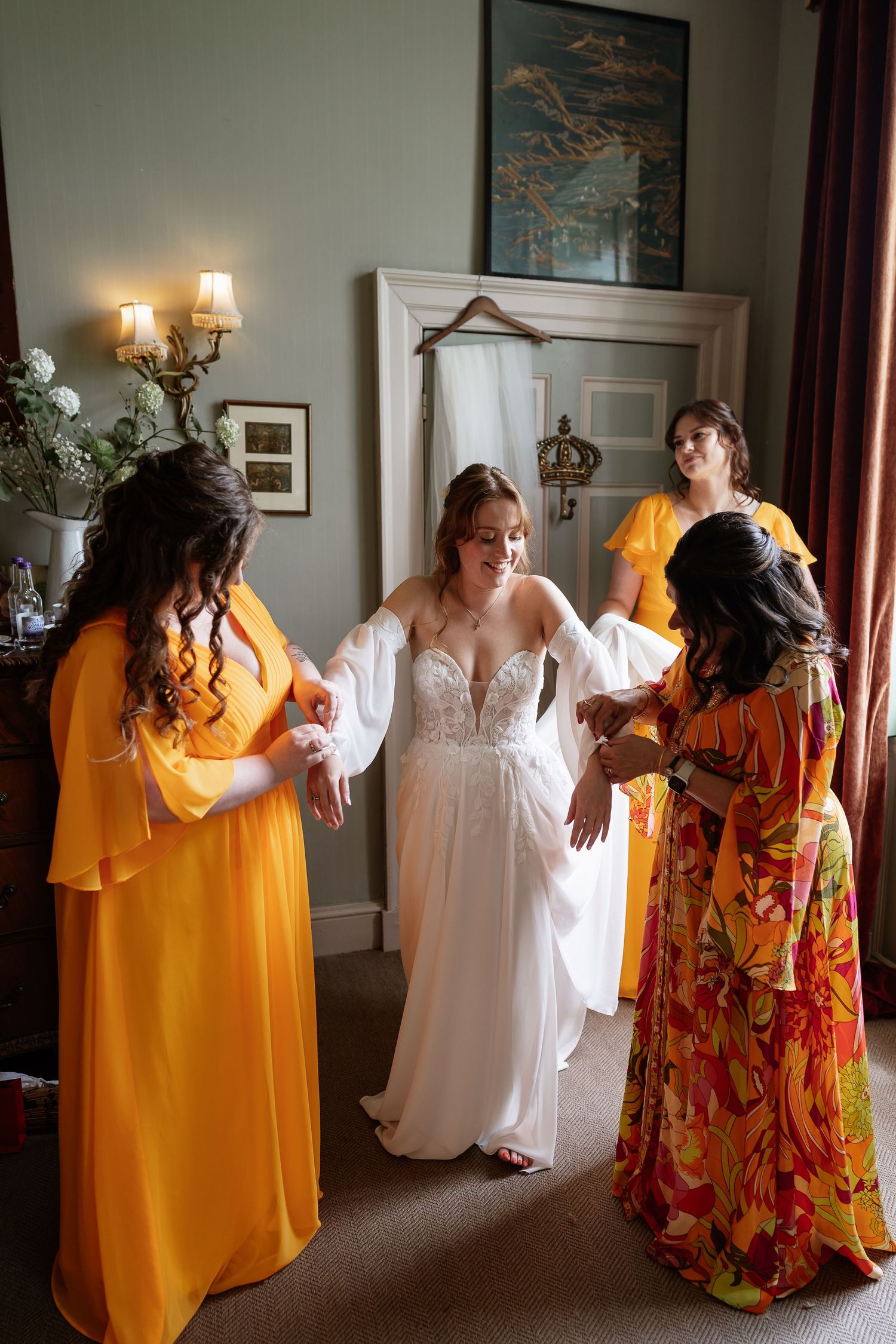 Bride in lace wedding dress being helped by bridesmaids in yellow gowns while getting ready.