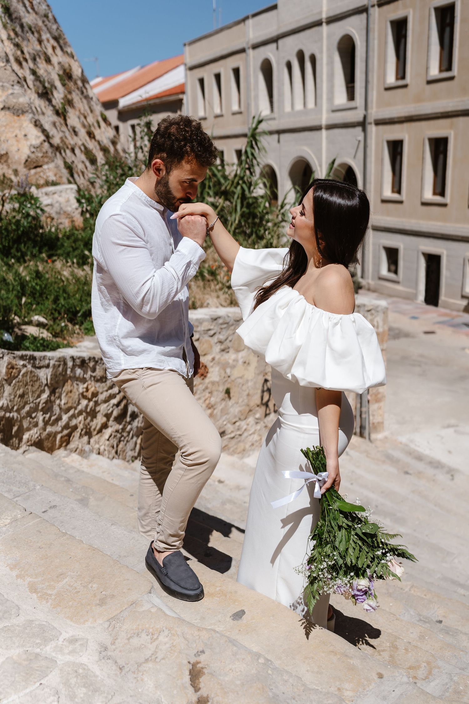 Groom kissing bride's hand on ancient stone steps with historic Mediterranean building in background.
