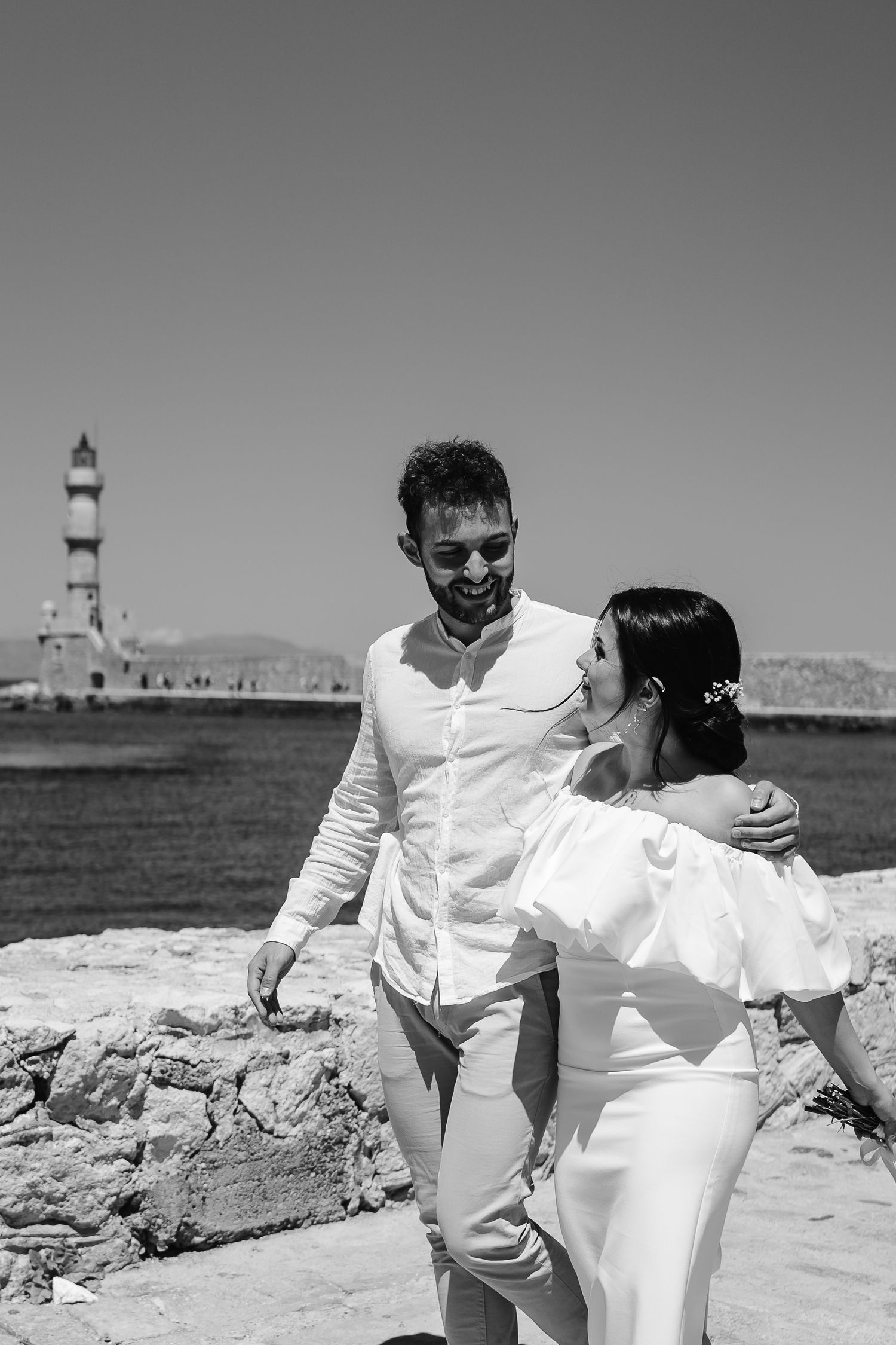 Black and white photo of laughing bride and groom walking near the Chania lighthouse in Crete.