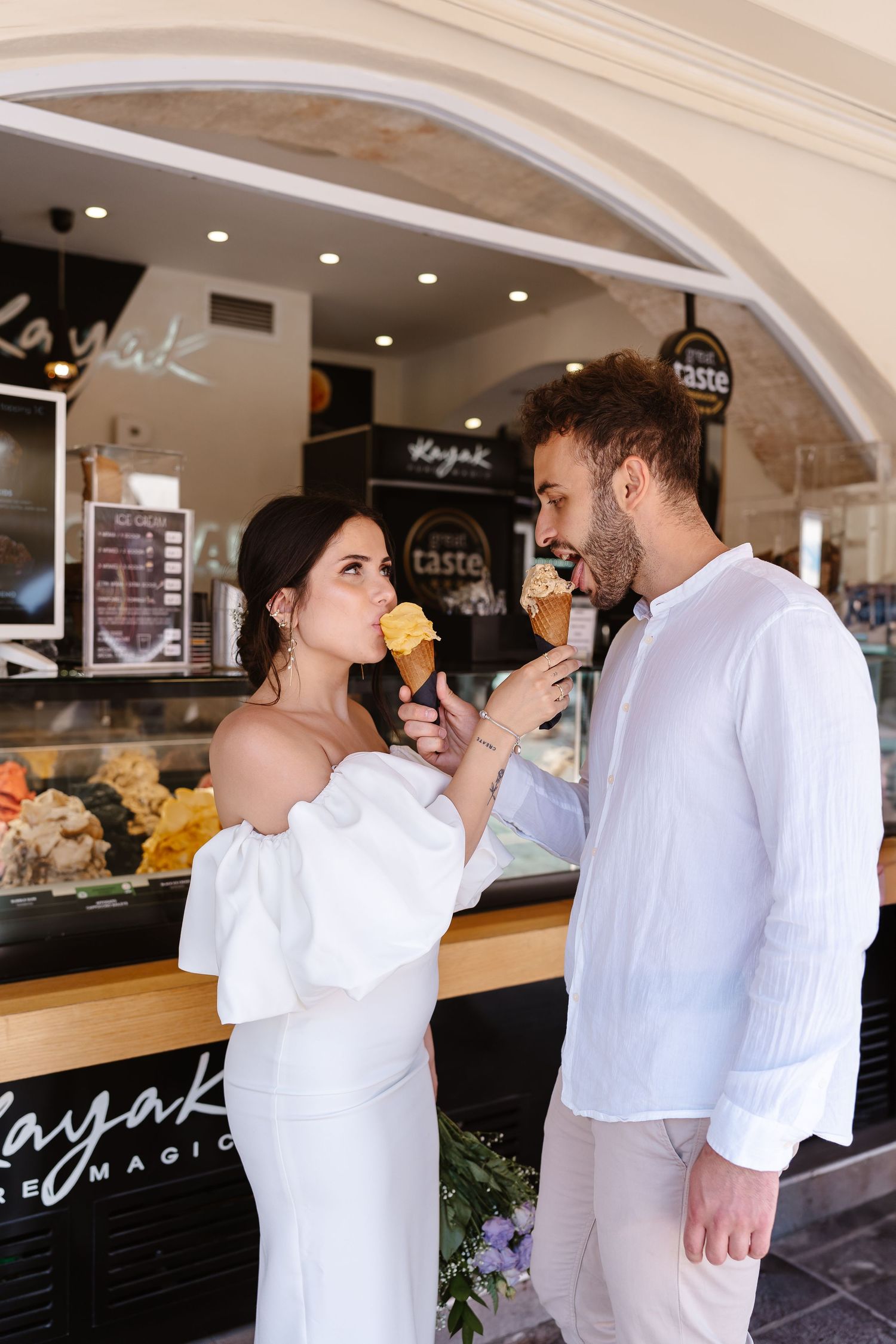 Bride in white off-shoulder dress feeds groom an ice cream cone outside a gelato shop.