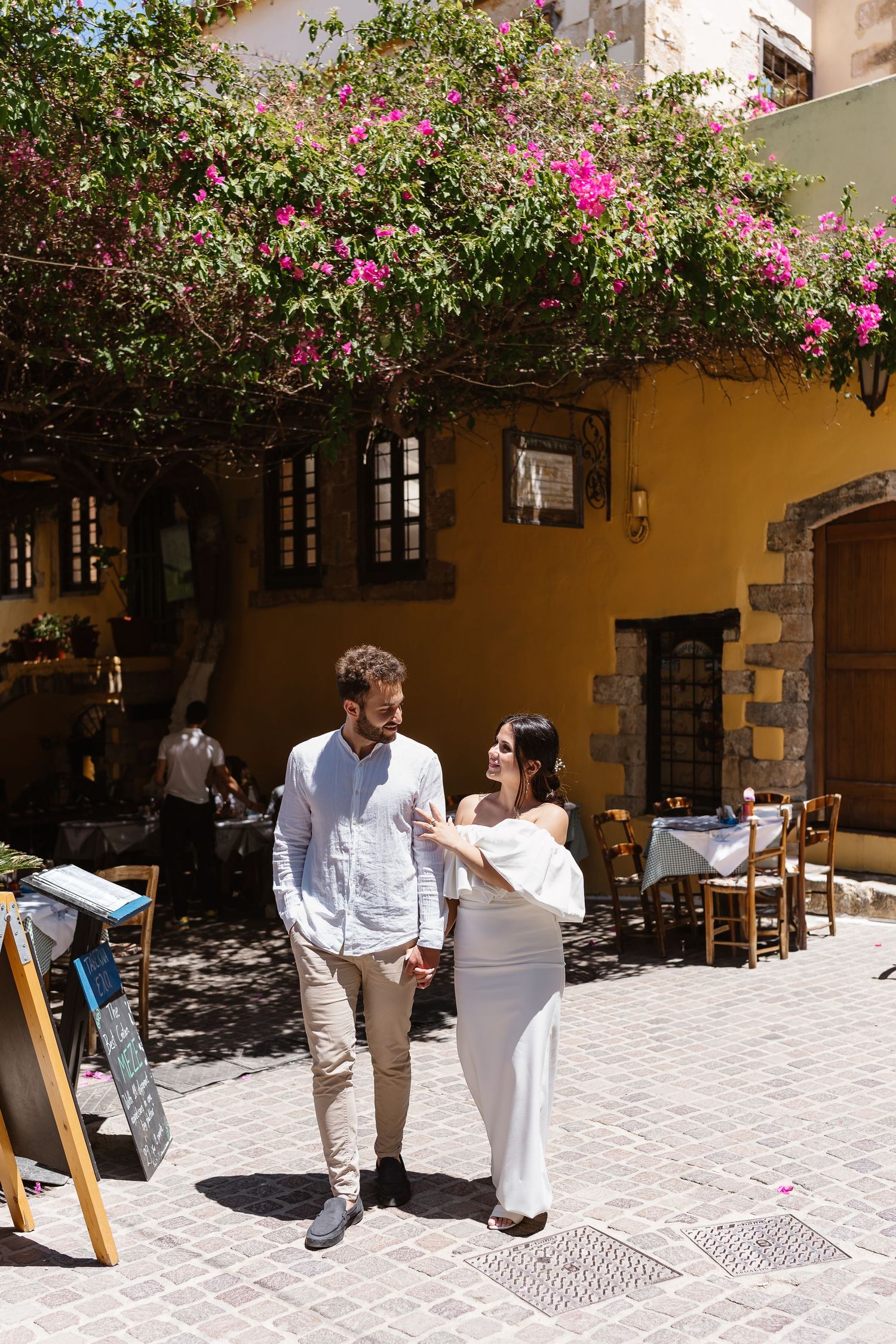 Couple walking hand in hand through sunny Mediterranean plaza, pink bougainvillea blooming above yellow building.