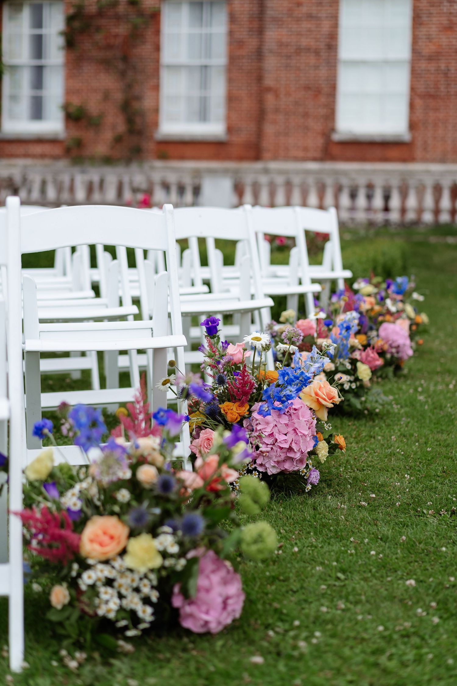 Colorful wildflower arrangements lining aisle of white chairs at outdoor wedding ceremony on green lawn.