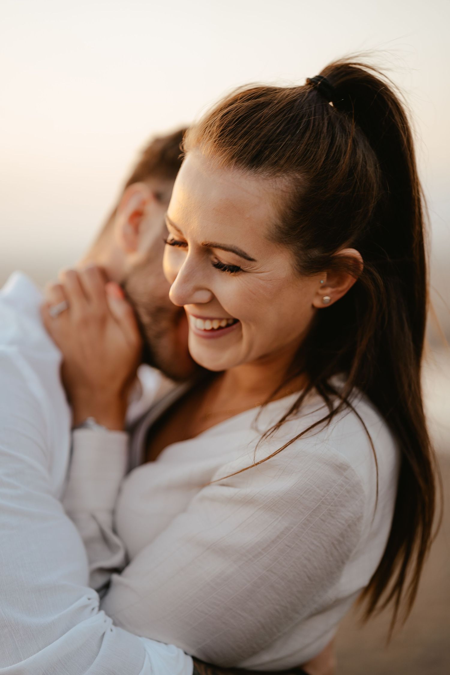 Close-up of smiling woman being embraced from behind by partner at sunset, wearing a light beige top.