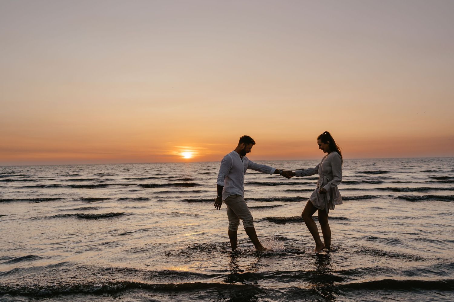 Romantic couple holding hands walking in shallow ocean water at sunset.