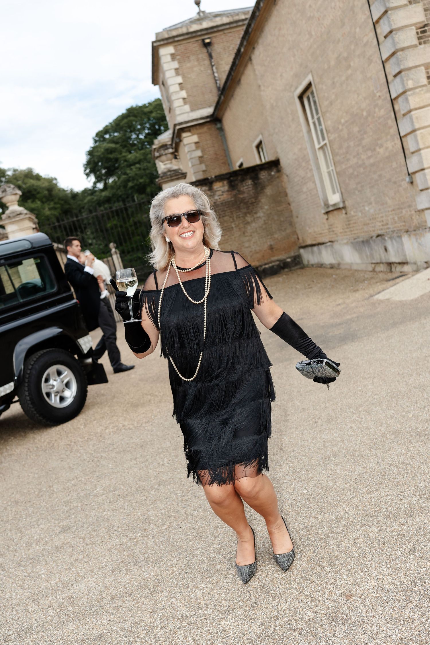 Woman in black fringe flapper dress with pearls and gloves walks outside historic stone building.