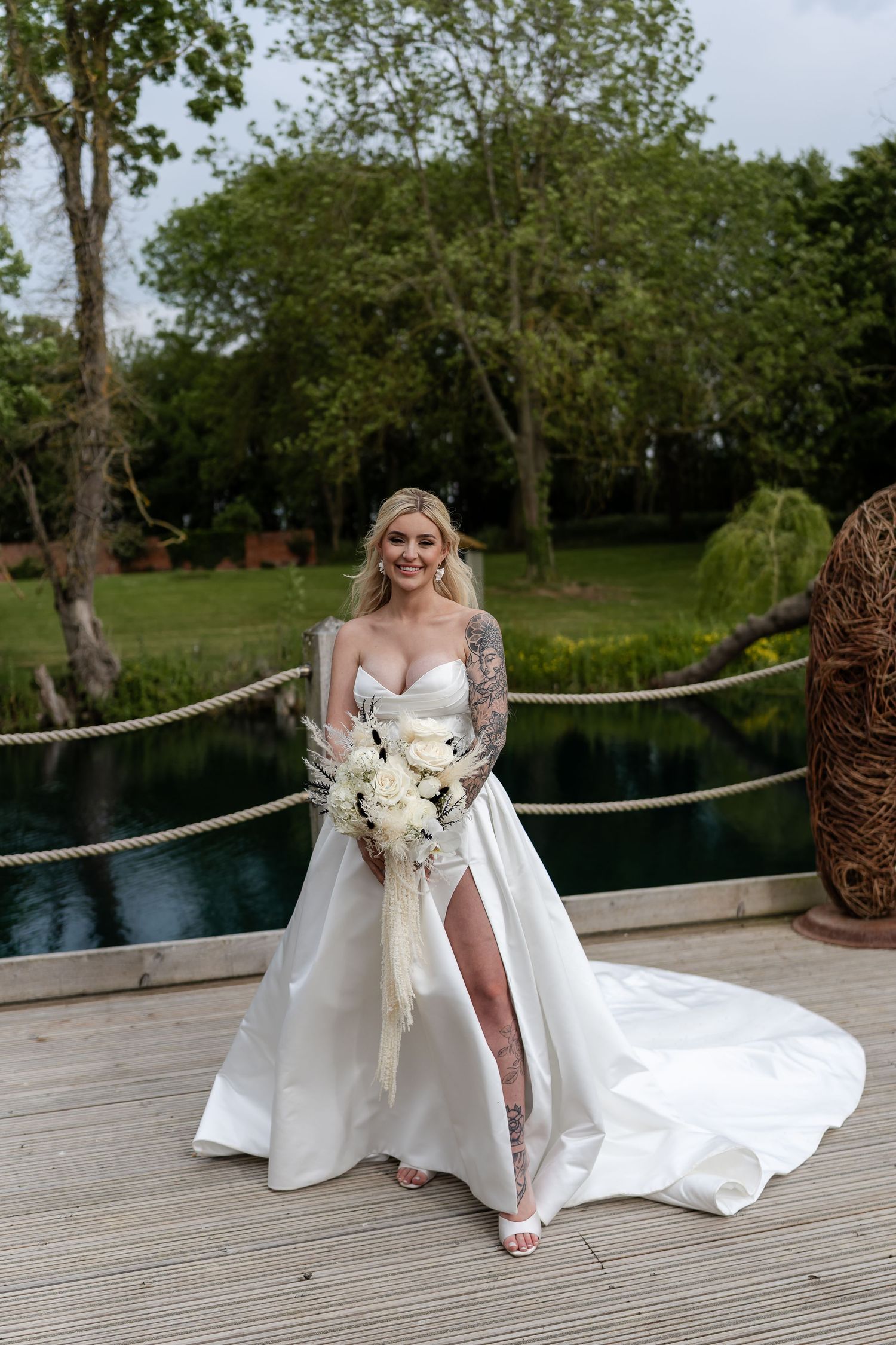 Elegant bride in a white satin gown with lace sleeves and thigh slit holding a bouquet on a lakeside deck.