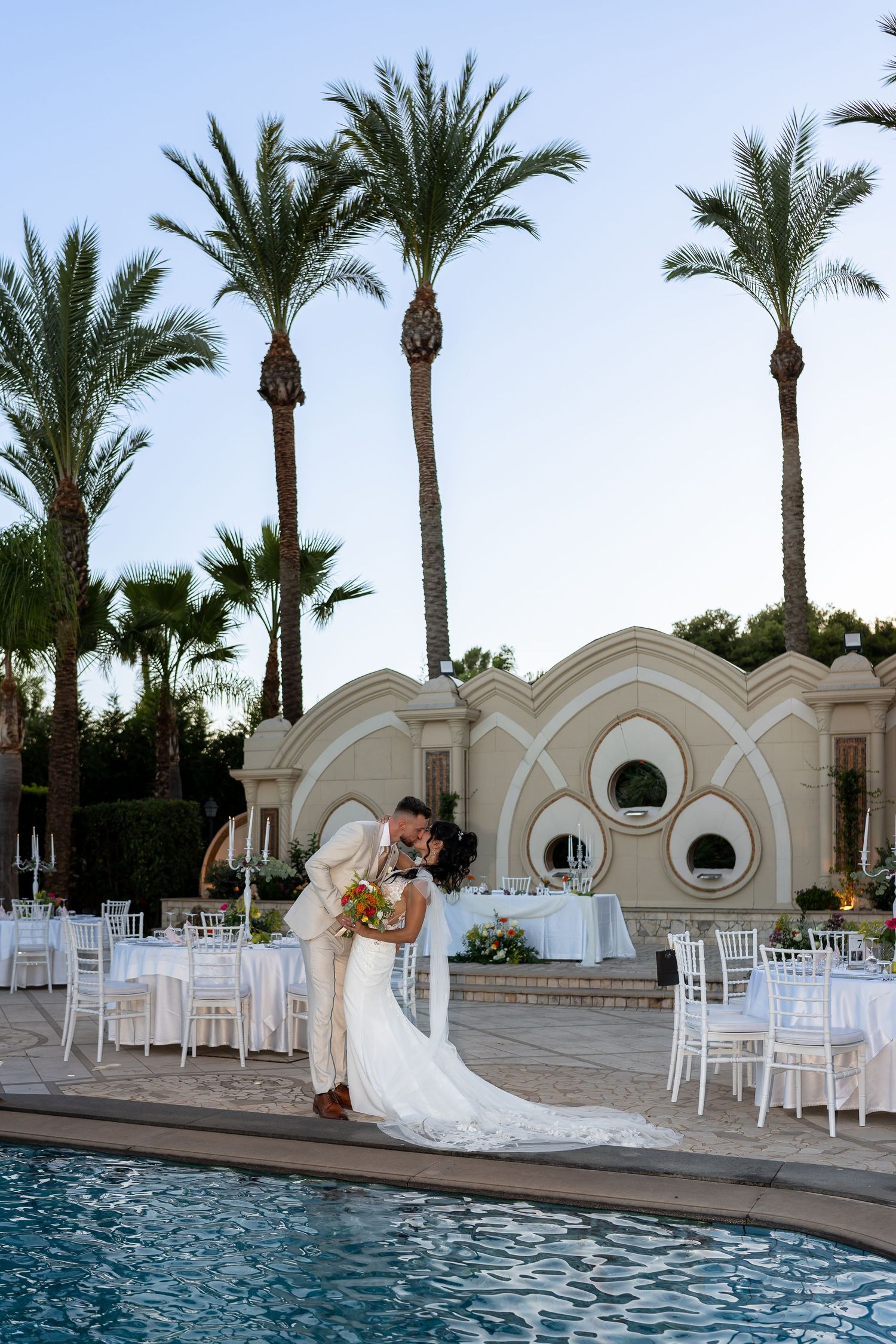 Bride and groom share a romantic dip kiss beside a pool with palm trees at an elegant resort venue.