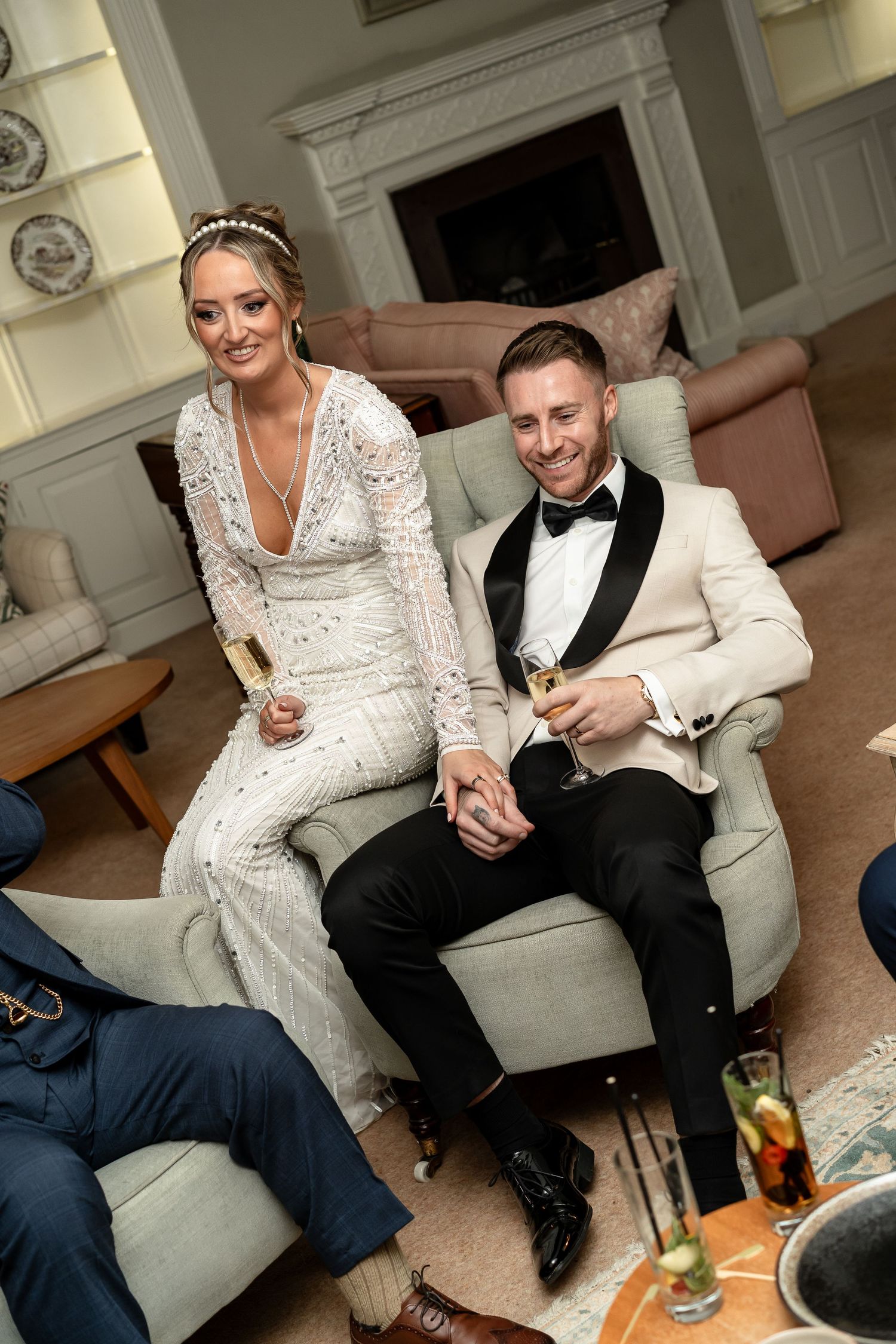 Smiling bride and groom relax on sofa with champagne glasses in elegant country house interior.