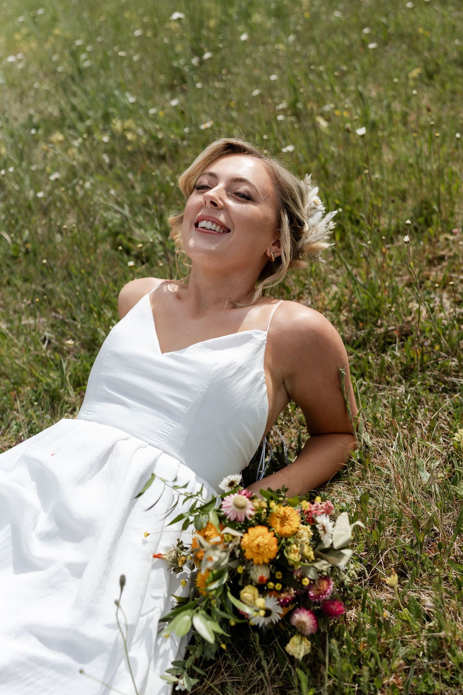 A joyful bride in a white dress lies in a wildflower meadow holding a colorful bouquet.