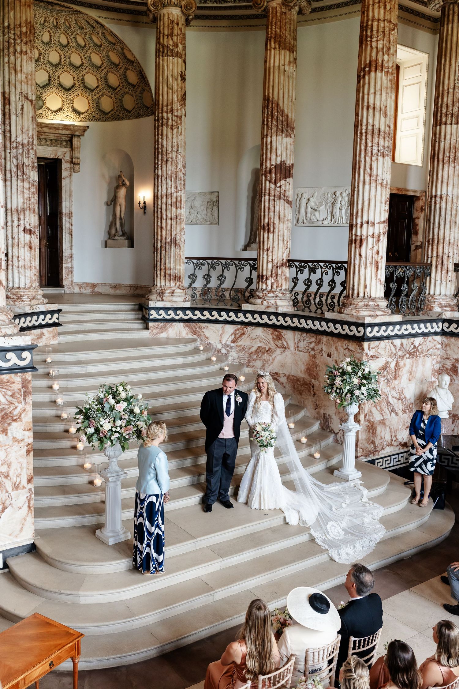 Bride and groom exchange vows on grand marble staircase in elegant neoclassical venue with floral arrangements.