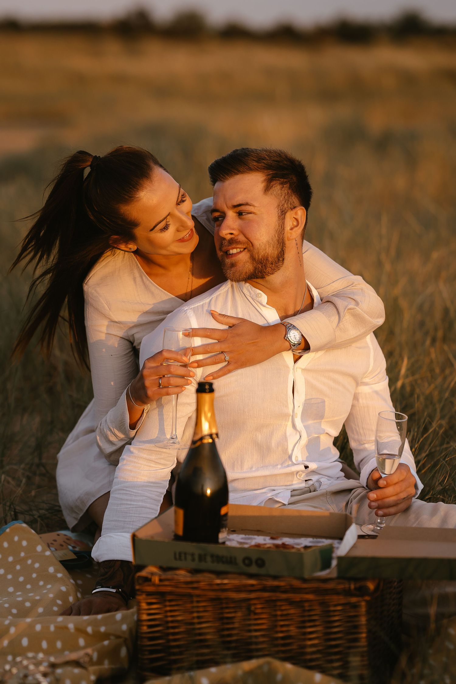 Couple enjoying a romantic champagne picnic in a golden field at sunset, woman embracing man from behind.