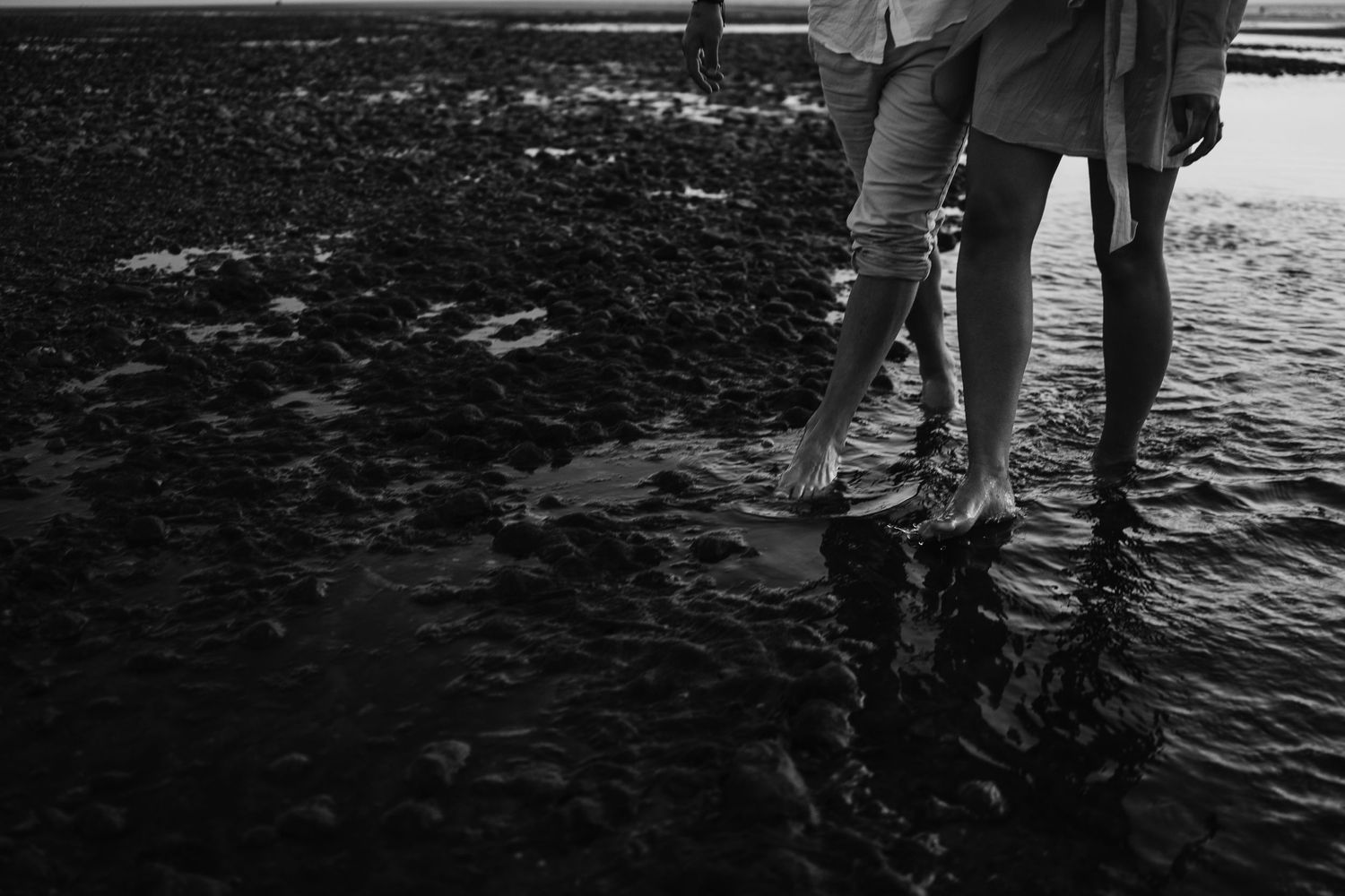 Black and white close-up of barefoot couple standing on rocky wet shoreline together.