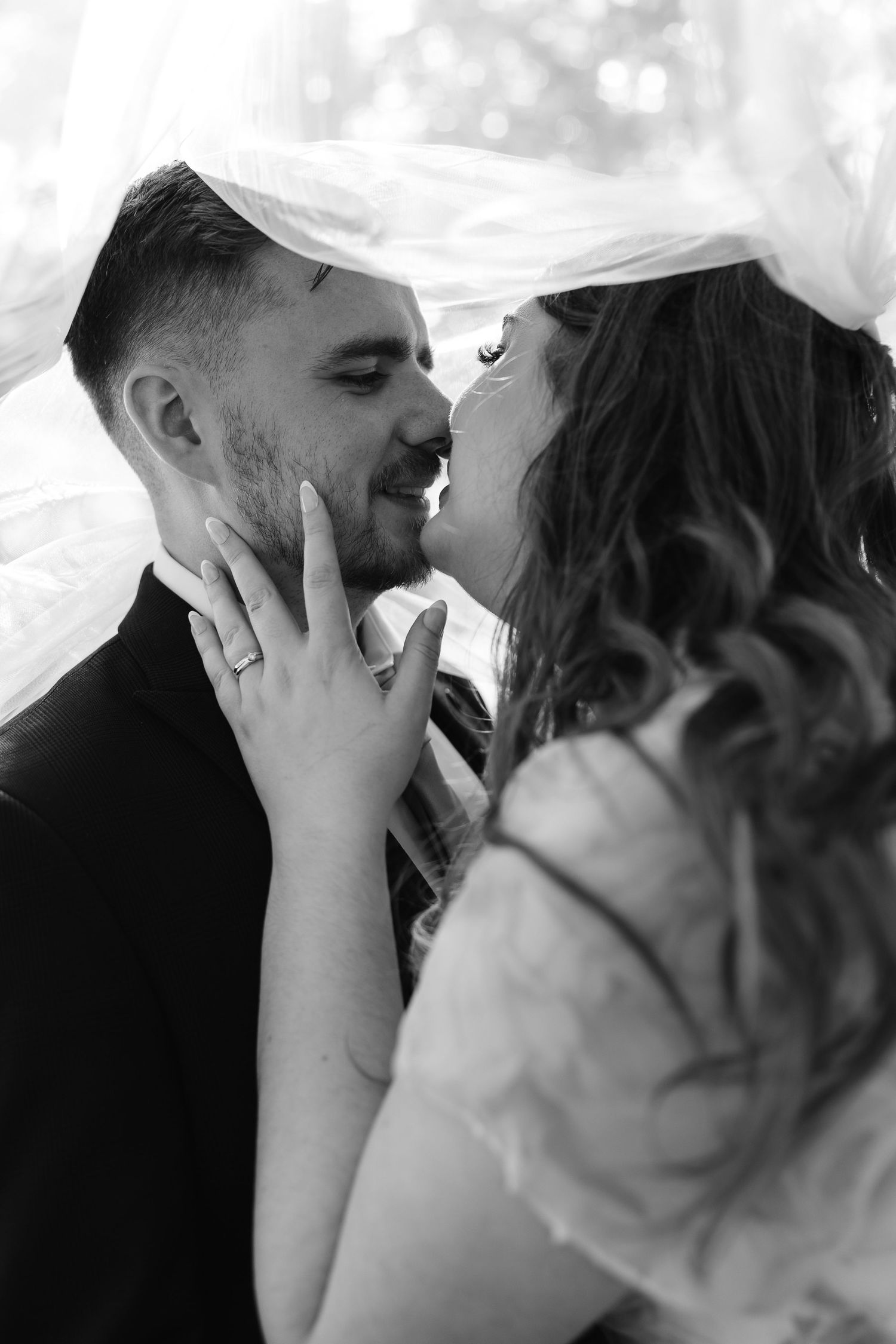 Intimate black and white portrait of bride and groom sharing a kiss beneath flowing wedding veil.