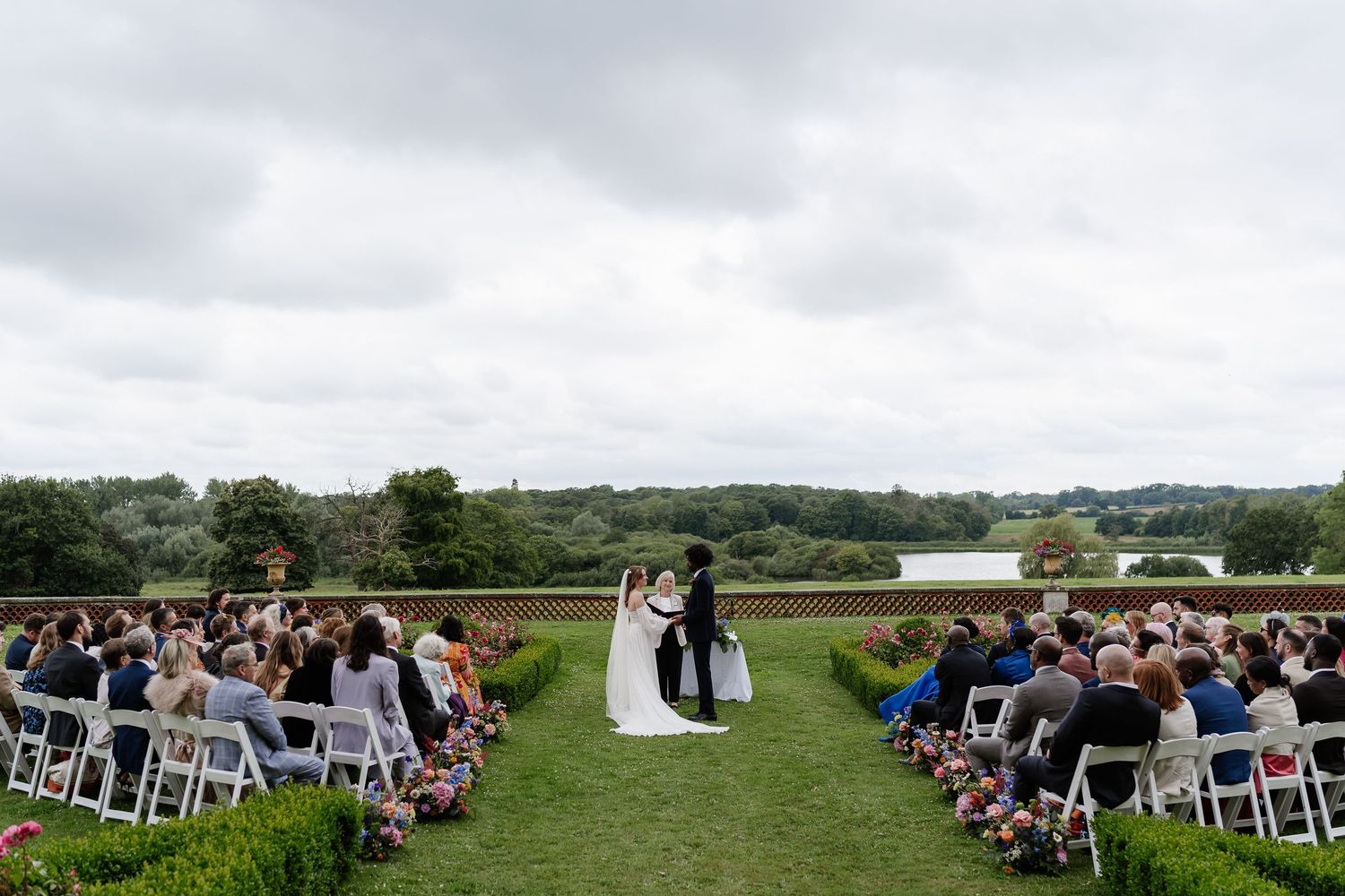 Wide shot of outdoor wedding ceremony with guests seated on white chairs overlooking scenic countryside lake.