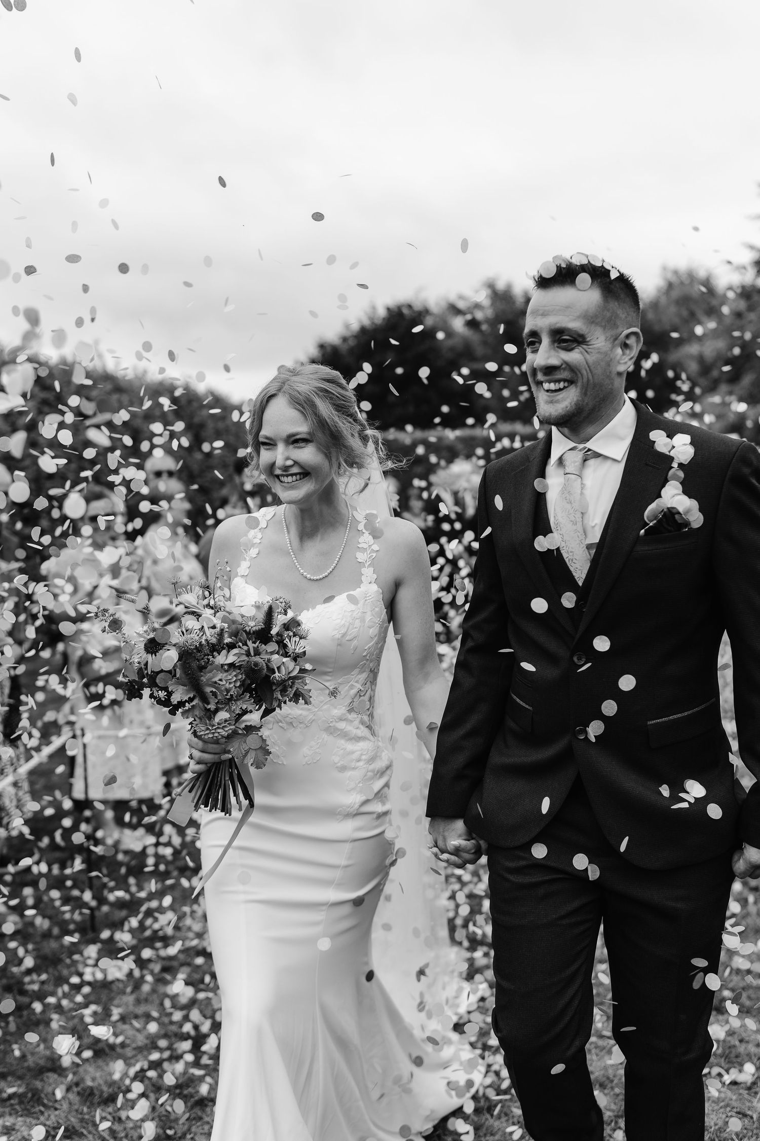 Smiling bride and groom walk through a shower of confetti in a joyful black and white wedding photo.