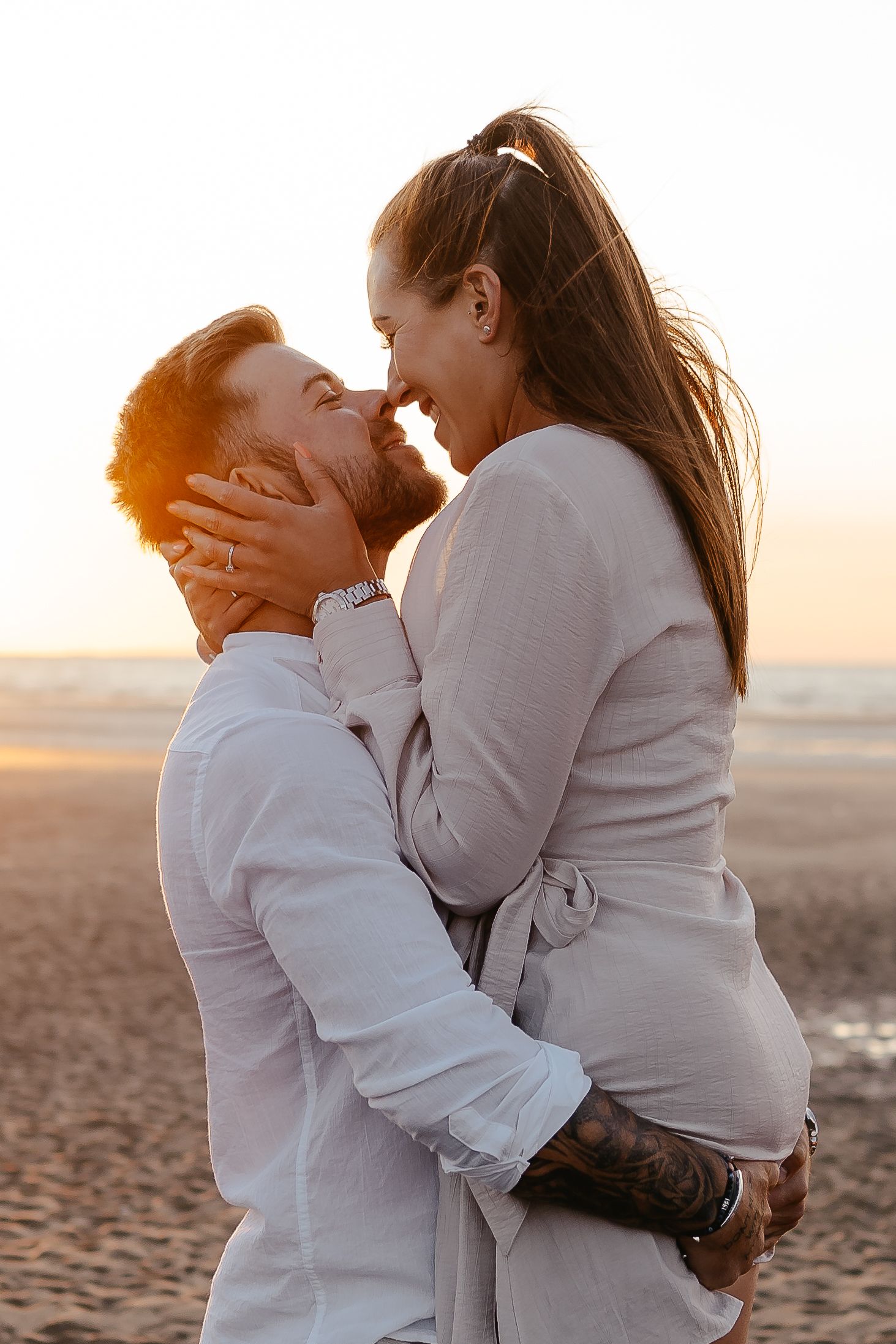 Couple sharing an intimate almost-kiss on a beach at sunset, woman held by man with tattoos.
