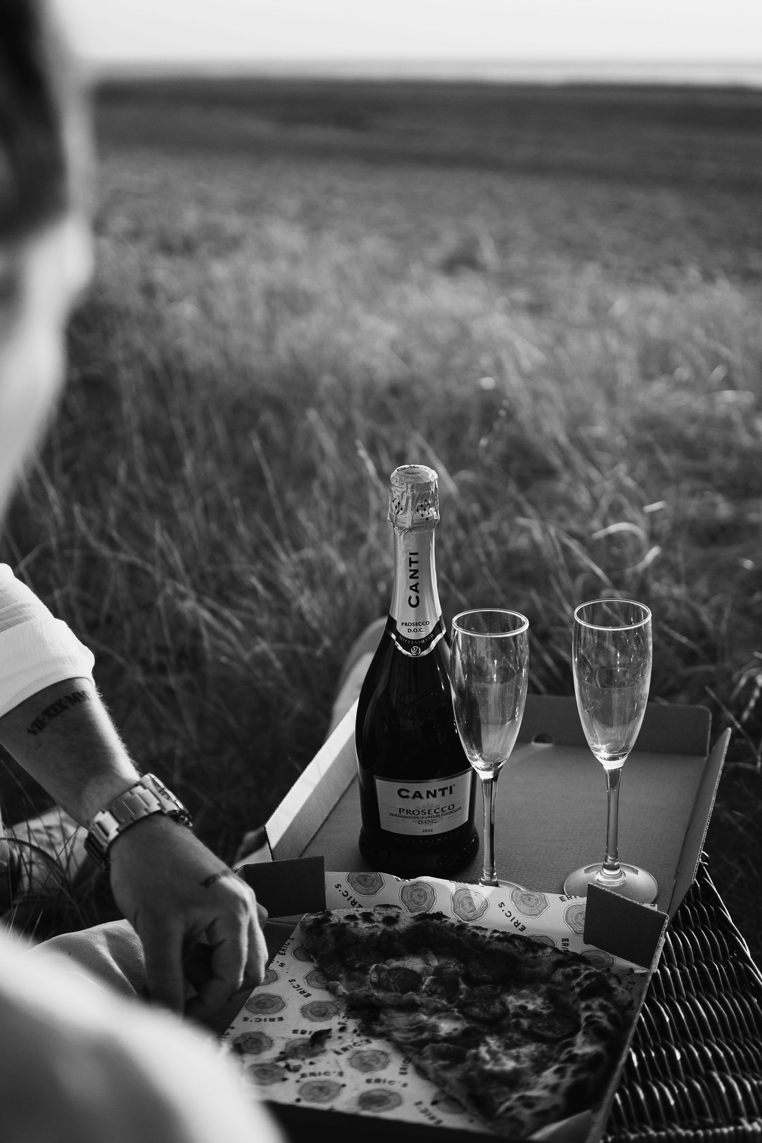 Black and white photo of a Canti champagne bottle with two flutes and pizza at an outdoor picnic.