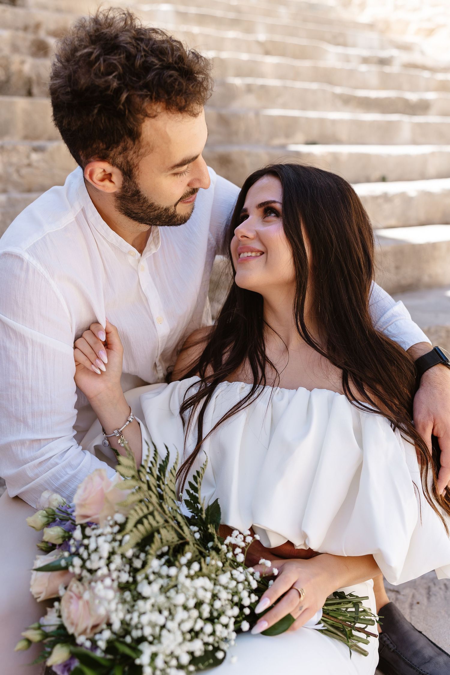 Happy couple sitting on ancient stone steps, groom embracing bride holding floral bouquet outdoors.