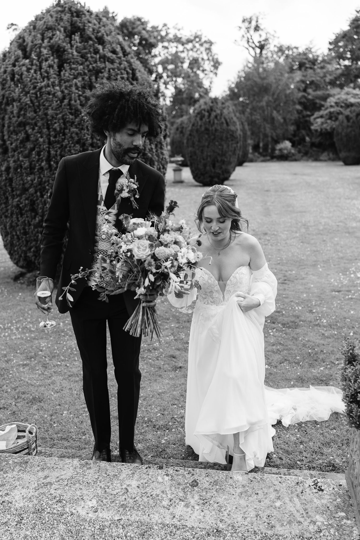 Black and white photo of newlyweds walking in garden, bride holding colorful bouquet.