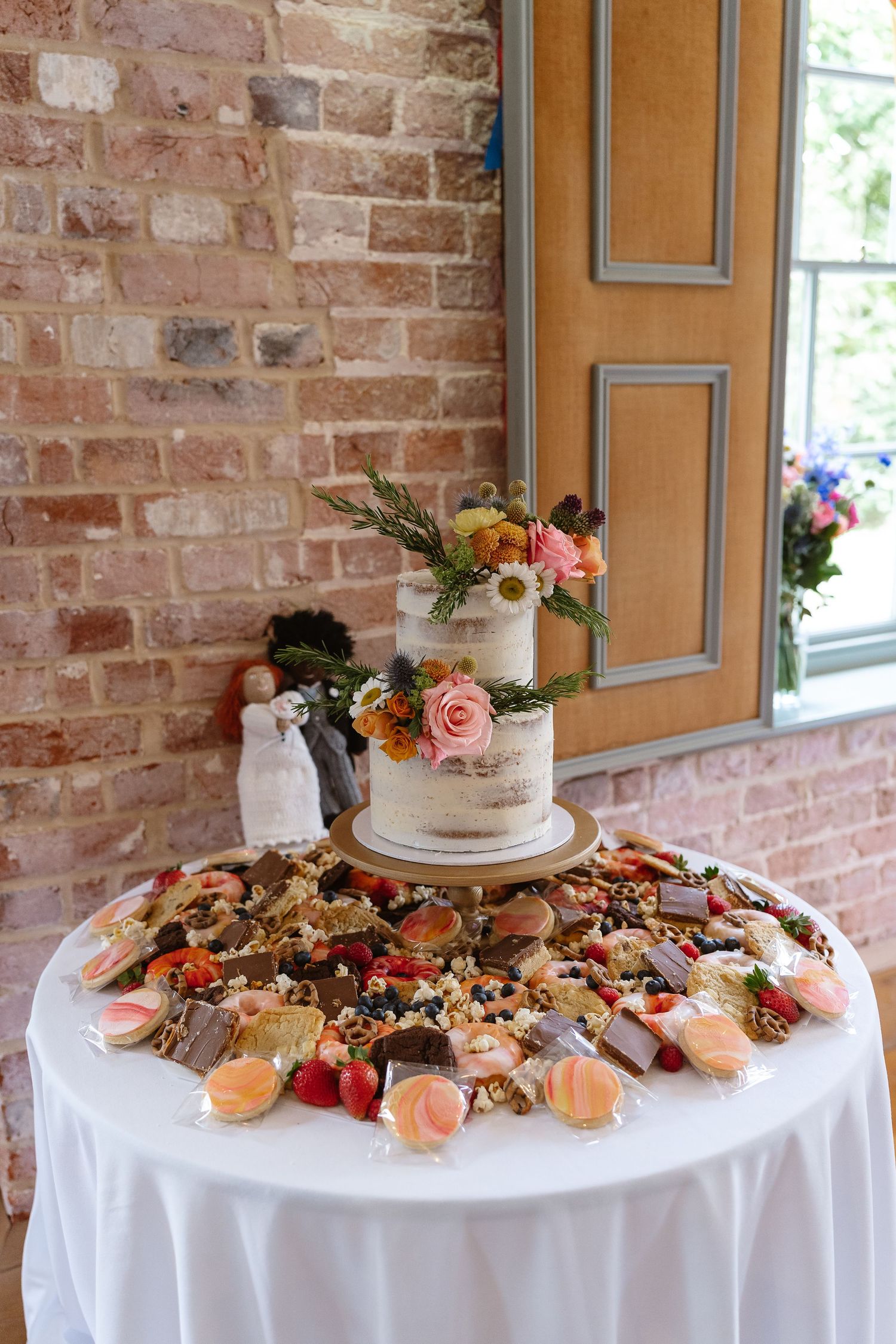Semi-naked wedding cake with floral topping surrounded by desserts and fruit on white table.