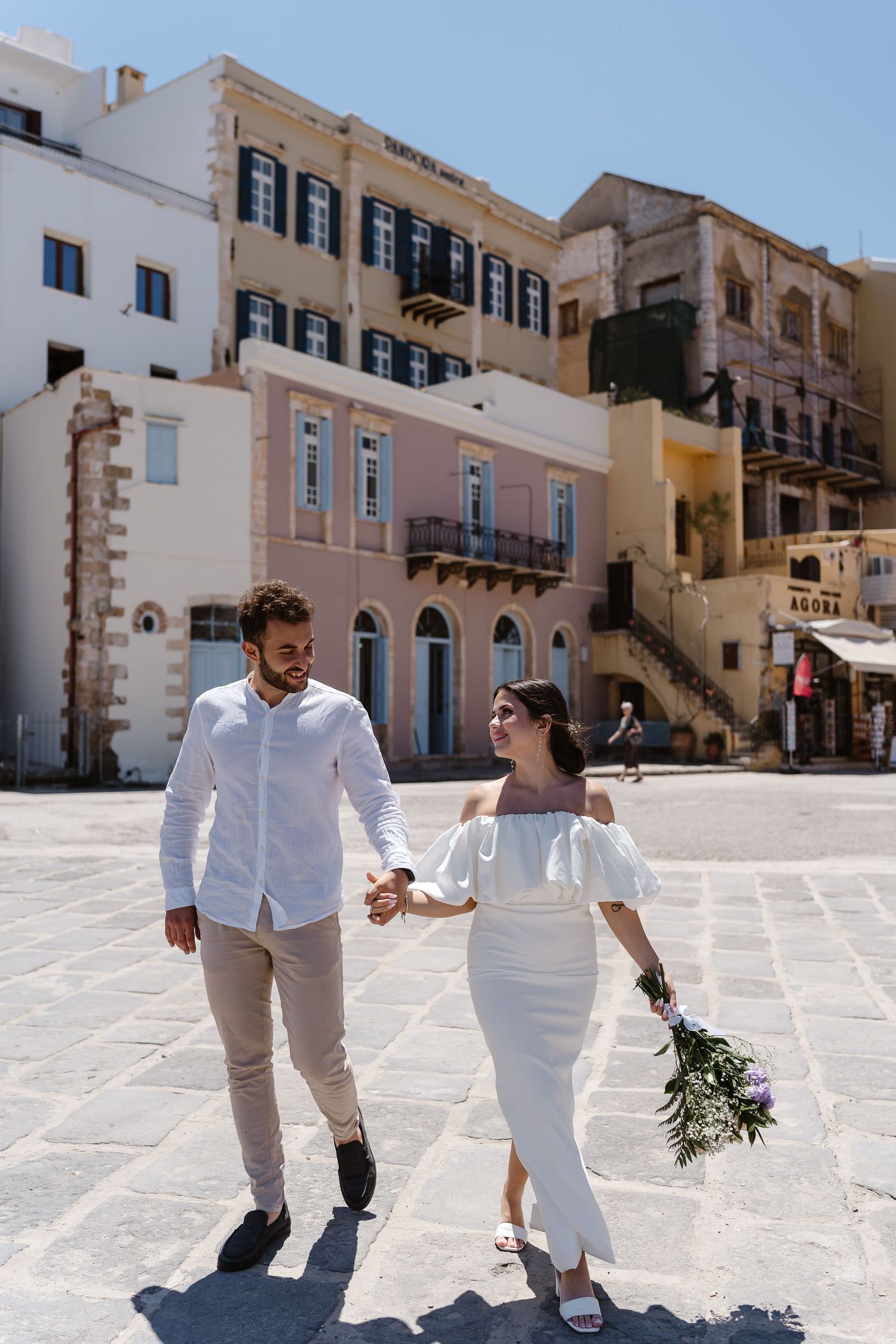 Smiling couple in white outfits holding hands walking across sunny Greek plaza with colorful historic buildings.