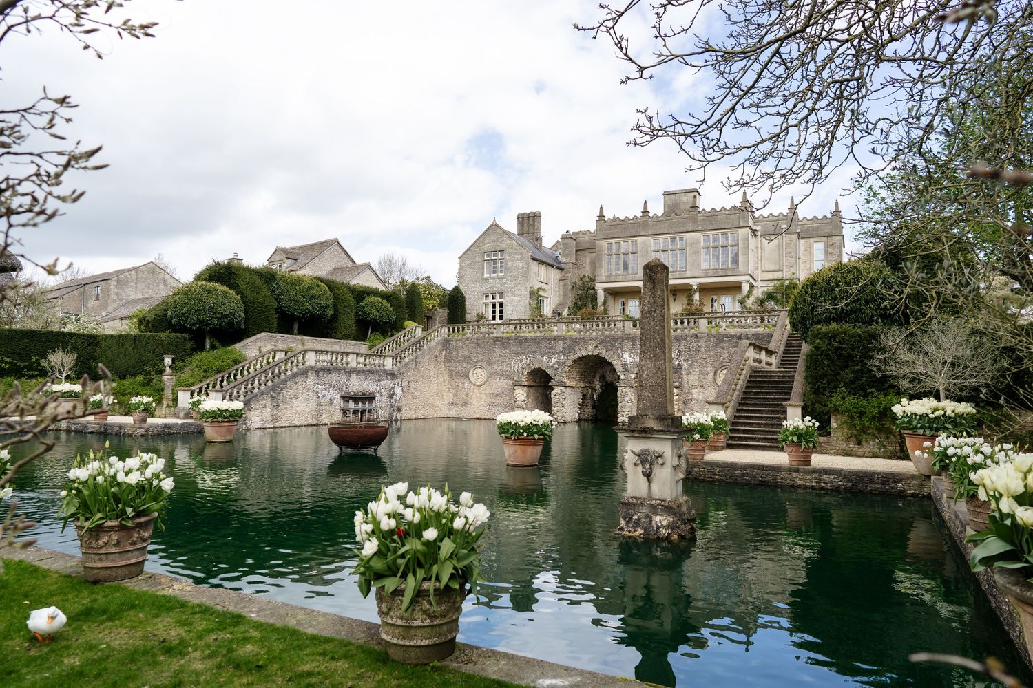 Elegant English manor wedding venue featuring ornate stone bridge, reflecting pool and white floral arrangements.