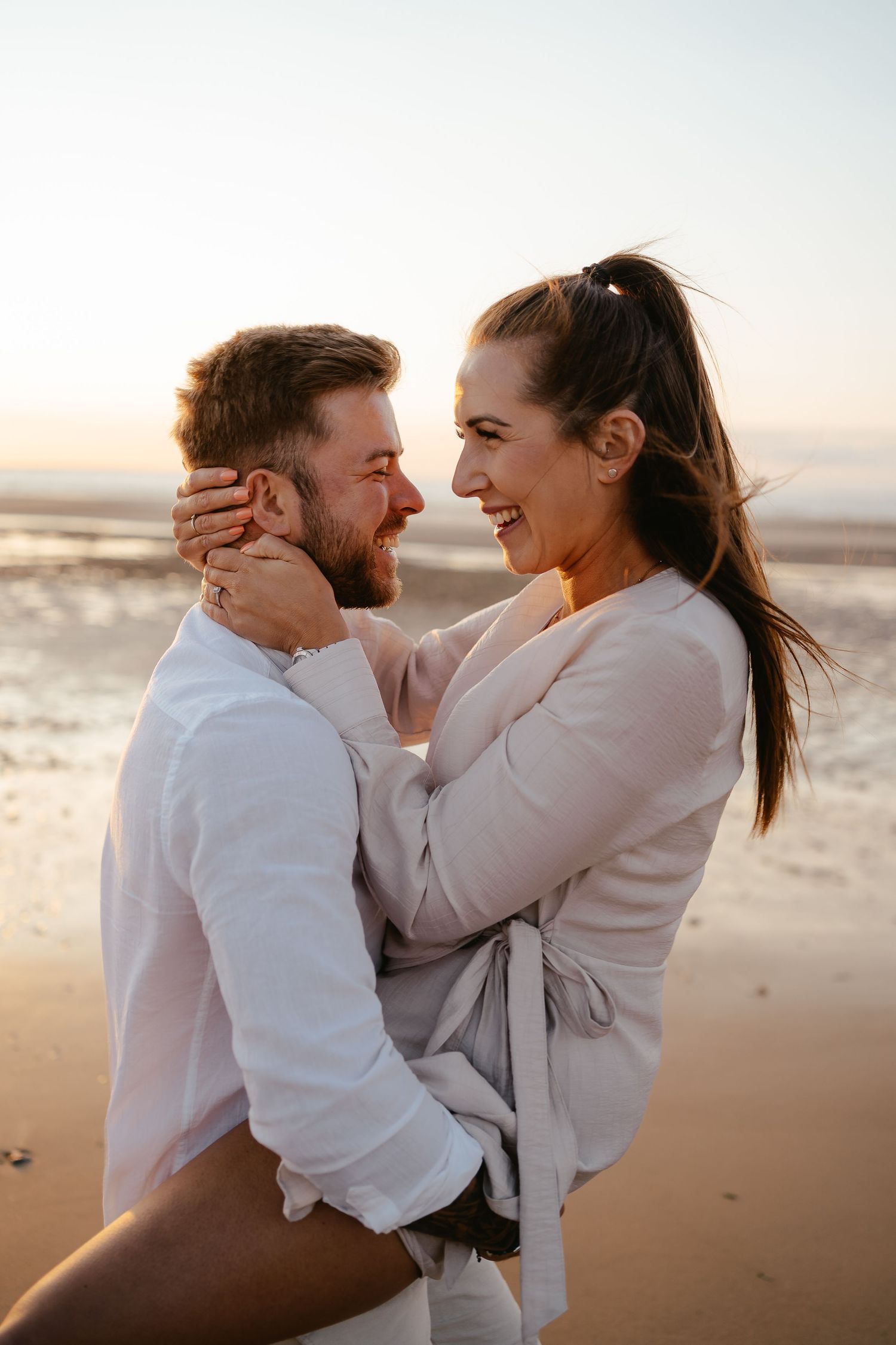 Couple sharing a romantic moment on a beach at golden hour, woman smiling while cradling mans face.