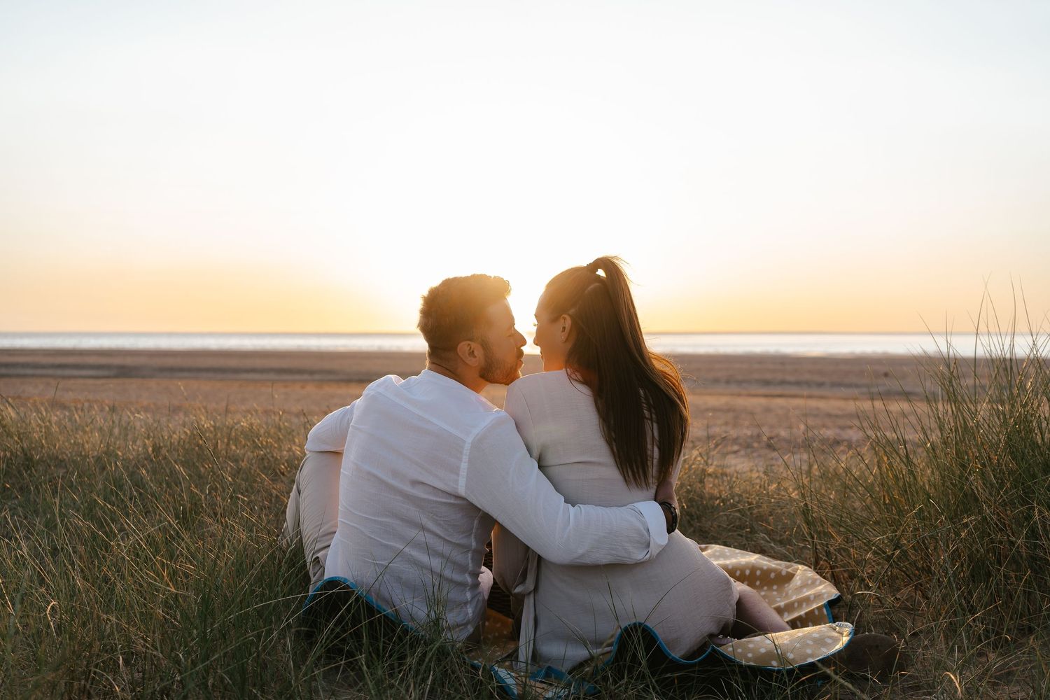 Couple sharing a tender kiss while sitting in beach grass during a golden sunset.