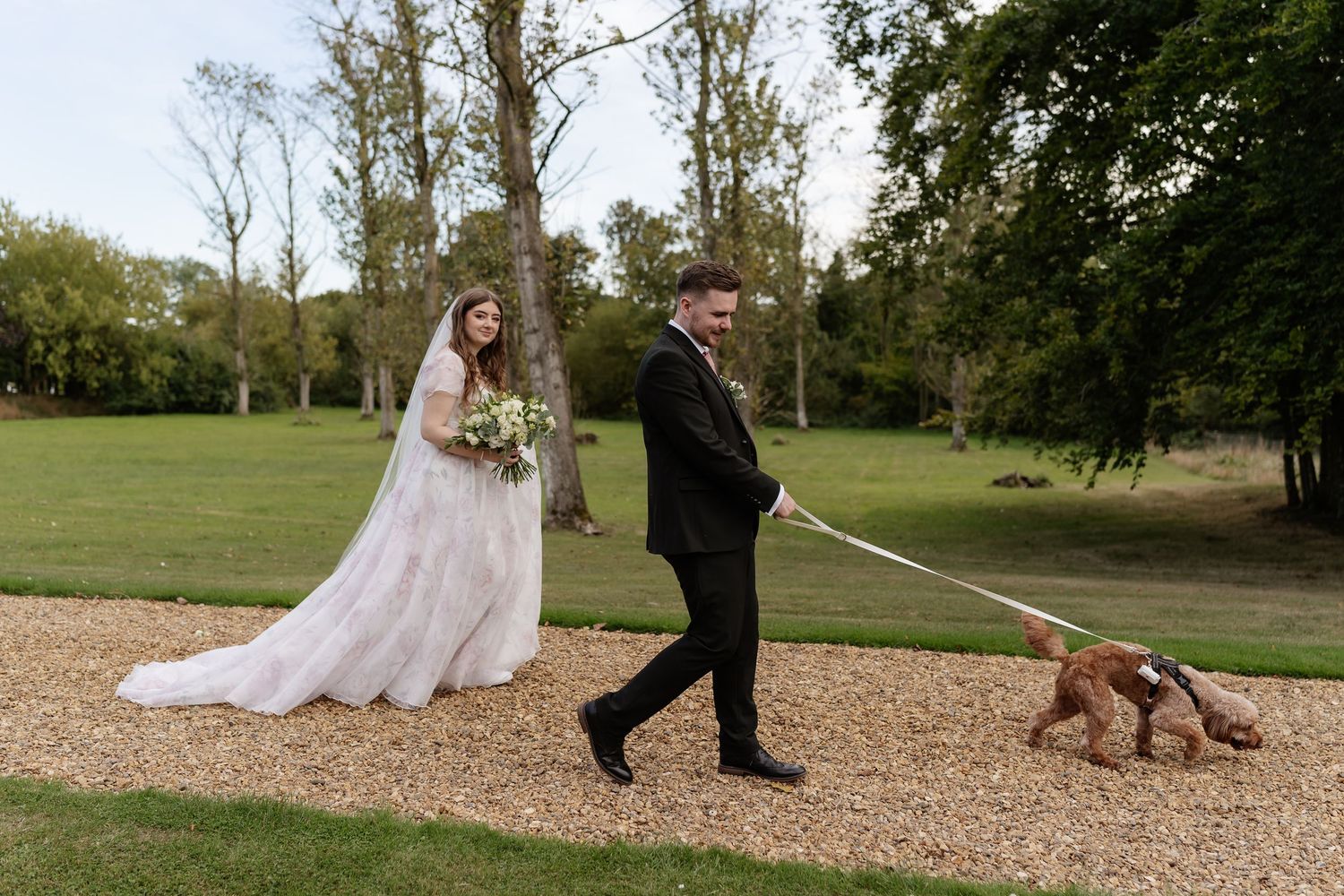 Bride and groom walking their golden dog on a leash along a gravel path at a country estate.