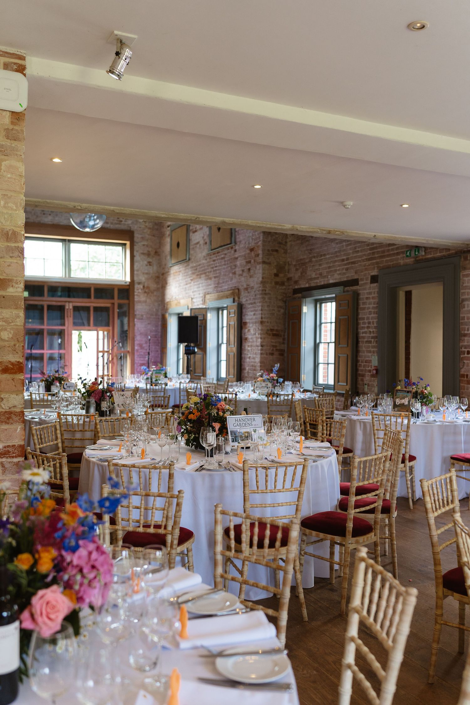 Elegant wedding reception hall with round tables, colorful floral centerpieces, and gold chiavari chairs.