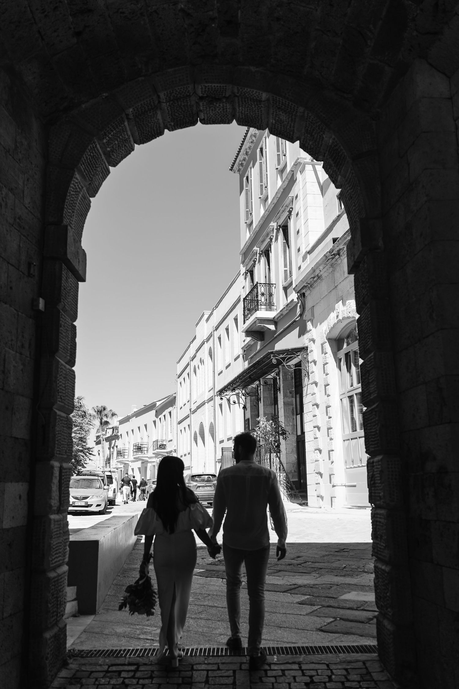 Black and white silhouette of couple holding hands walking through historic stone arch gateway.