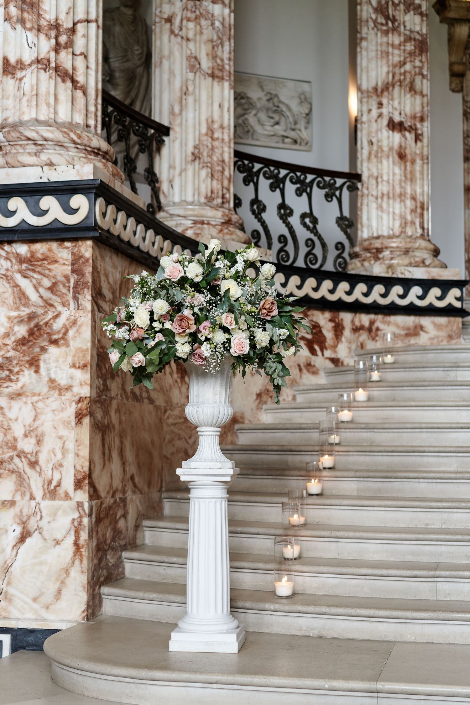 Elegant marble staircase with wrought iron railings, candles on steps, and a white pedestal floral arrangement with roses.