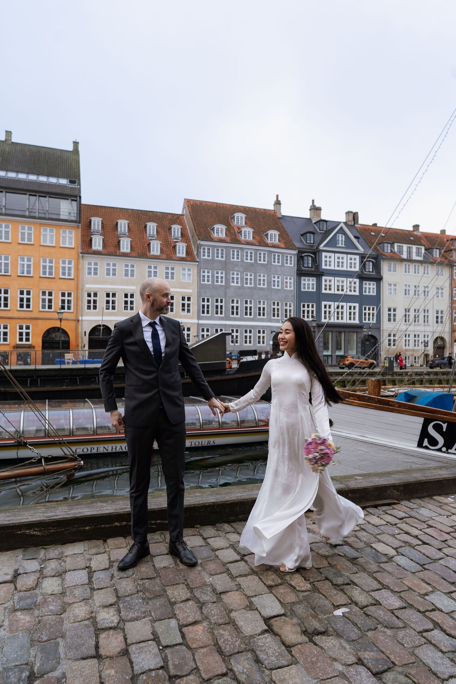 Bride and groom hold hands walking on cobblestone street at Nyhavn harbor in Copenhagen, Denmark.
