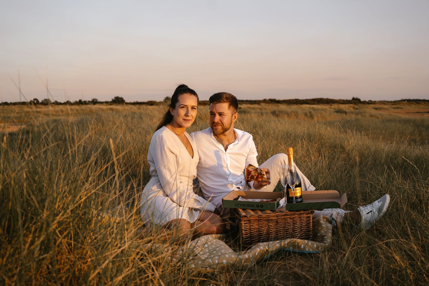 Couple sitting together in a golden grass field at dusk with a picnic basket.