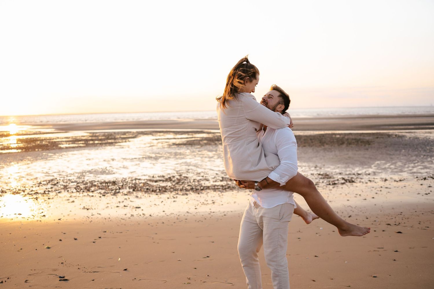 Man lifting woman in arms on sunny beach at sunset, both wearing white.